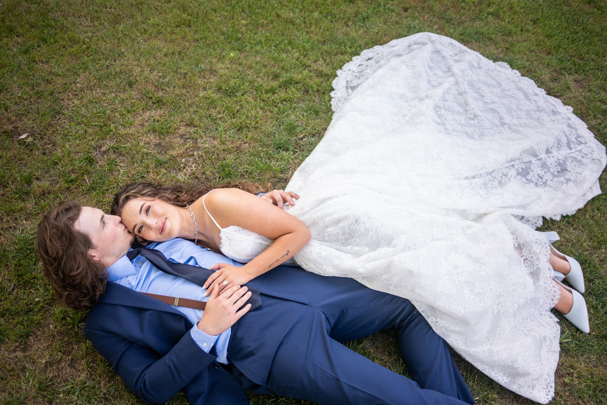 A bride and groom lie on grass, with the bride in a white wedding dress and the groom in a blue suit, smiling at each other.