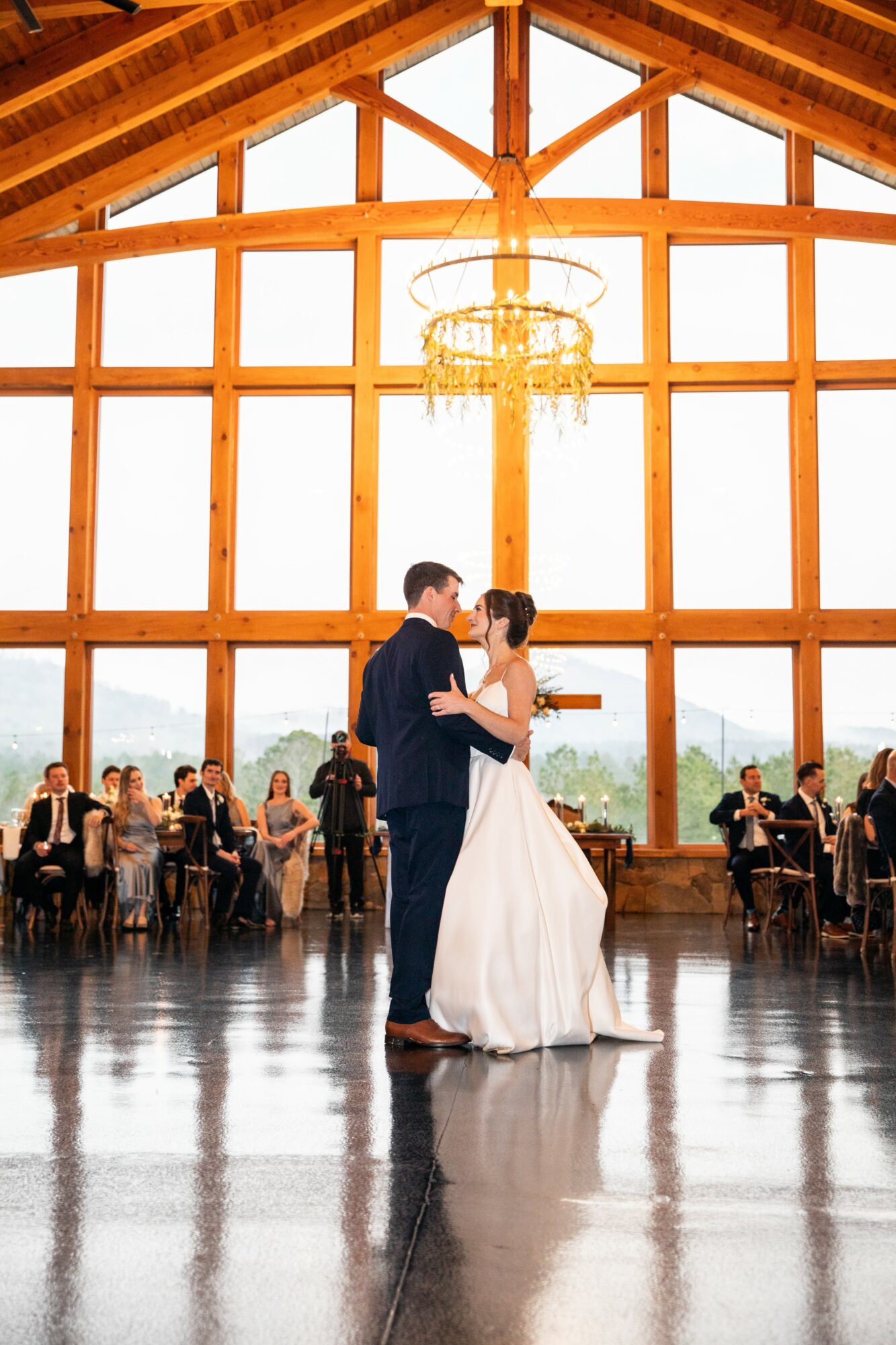 Couple dancing in a large wooden hall with high windows and a chandelier, surrounded by seated guests at tables.