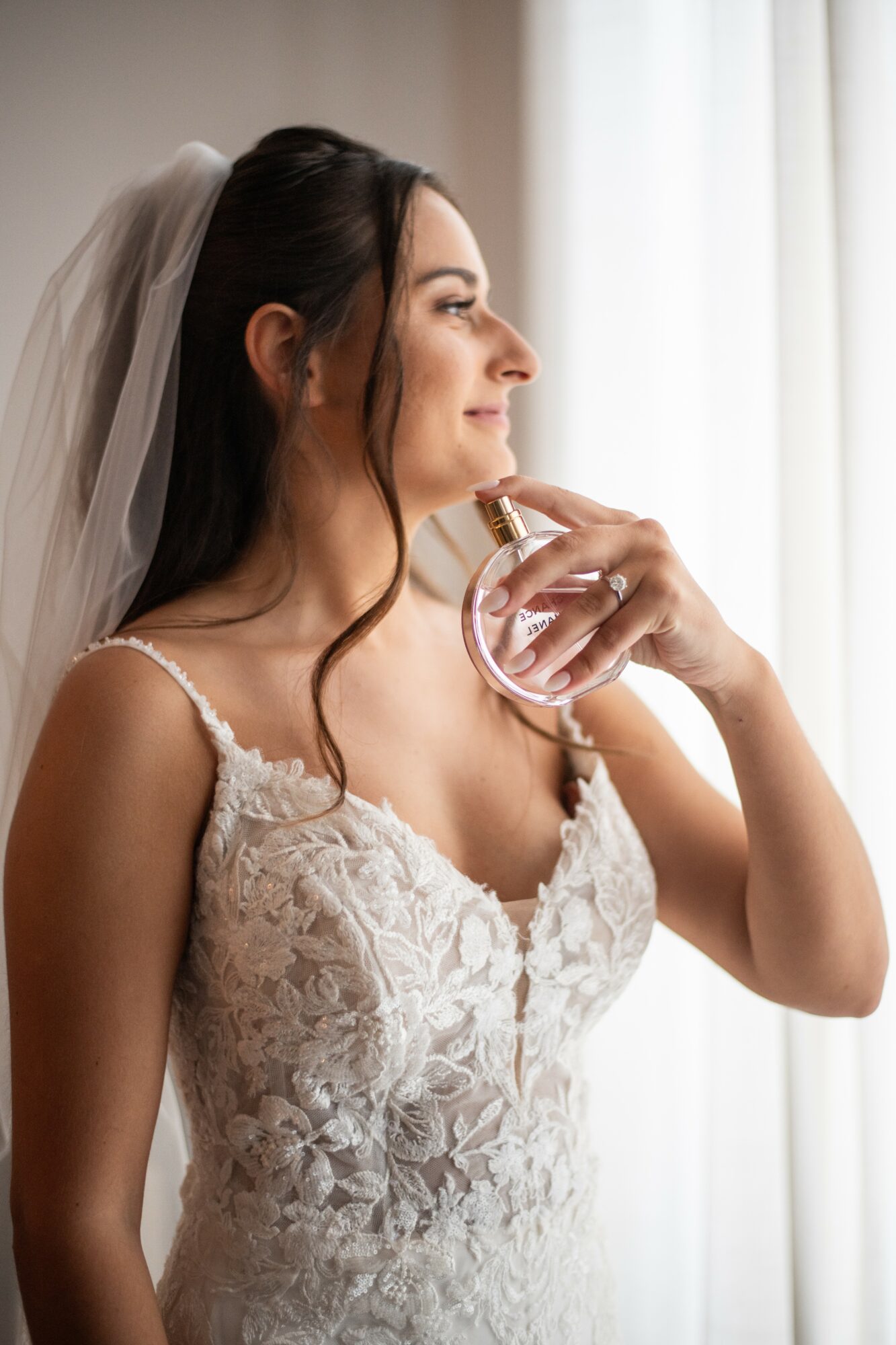 Bride in wedding dress and veil holding perfume bottle near her face, standing by window with white curtains, smiling softly.
