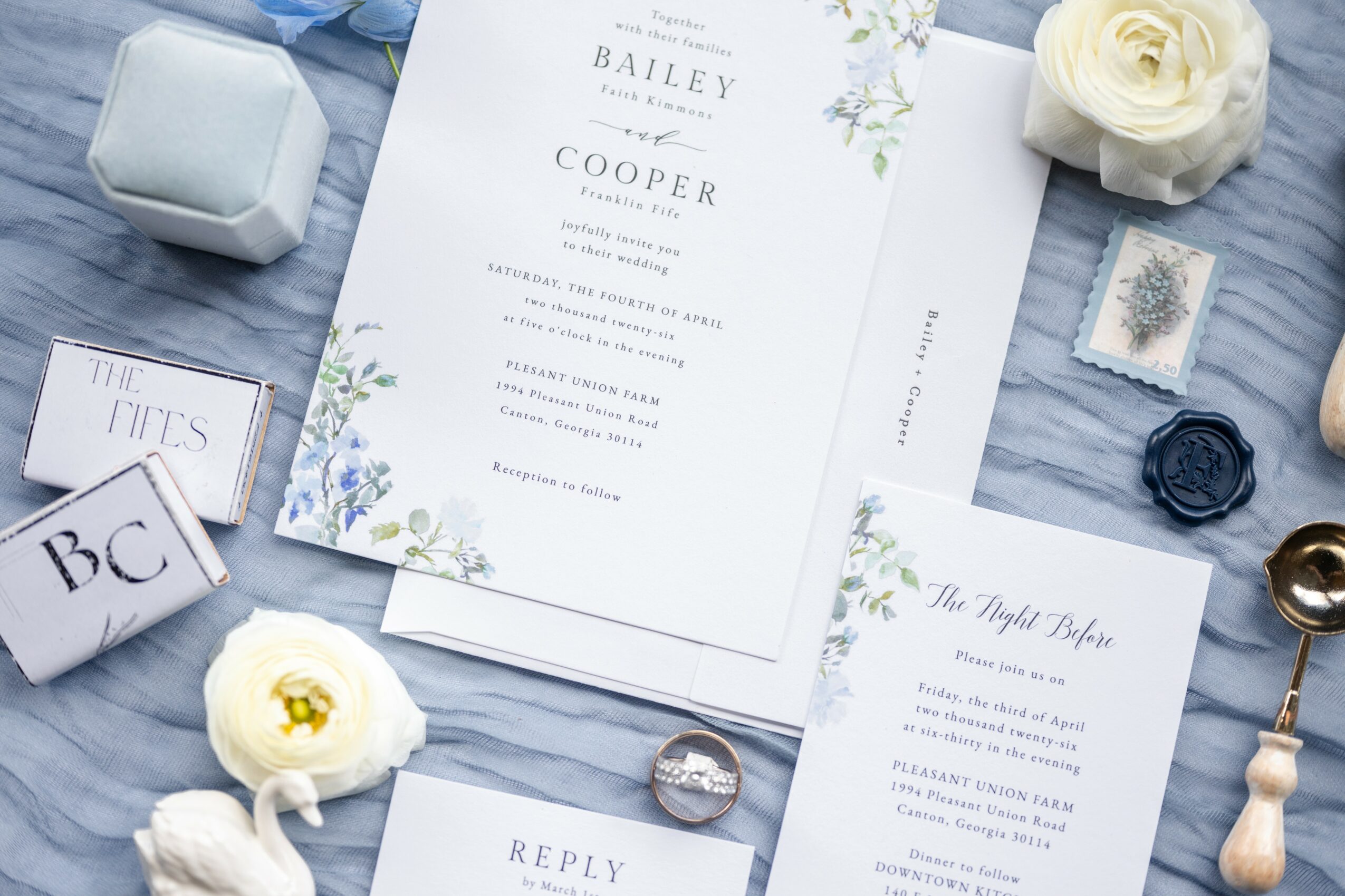 Wedding invitation with floral design, surrounded by flowers, stamps, and decorative items on a blue fabric background.