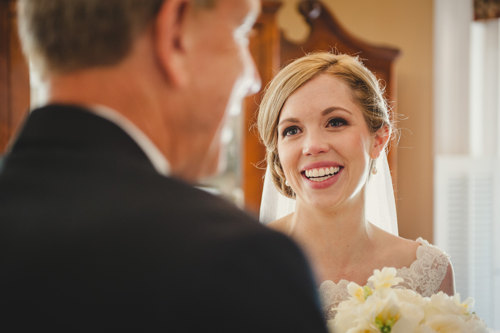 A smiling bride with blonde hair and a lace wedding dress looks at a man in a suit, indoors with warm lighting.