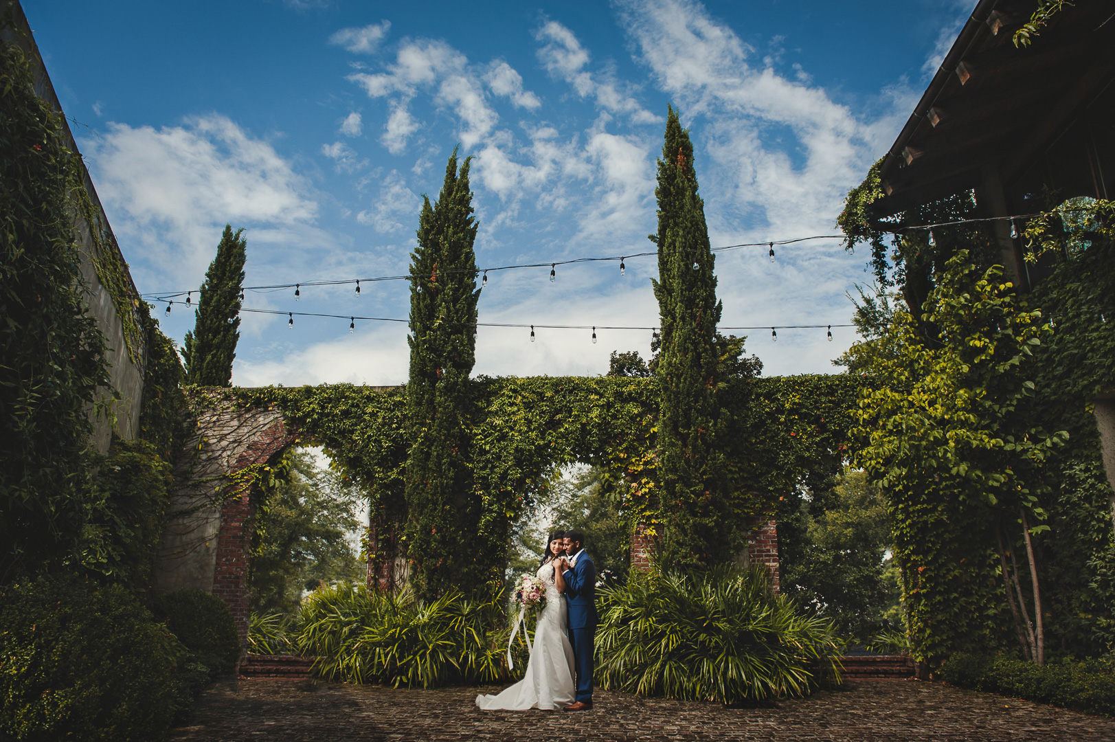 A couple in wedding attire standing outdoors with tall trees and a stone wall behind them, under a blue sky with clouds.