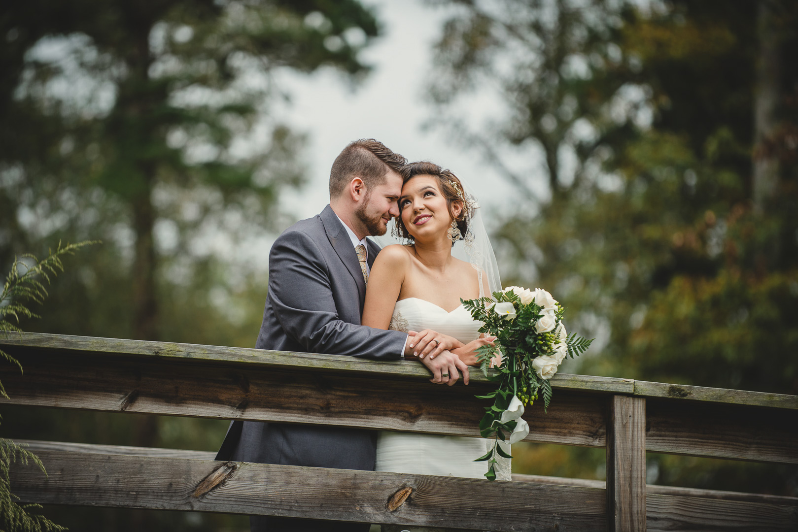 A bride and groom leaning on a wooden railing outdoors, smiling at each other, surrounded by trees.