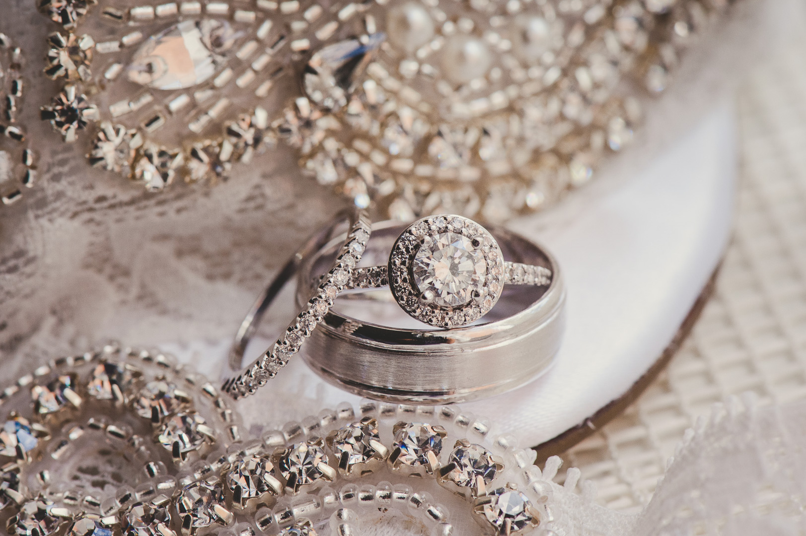 Close-up of a diamond engagement ring and wedding band on lace fabric with jewelry pieces nearby.