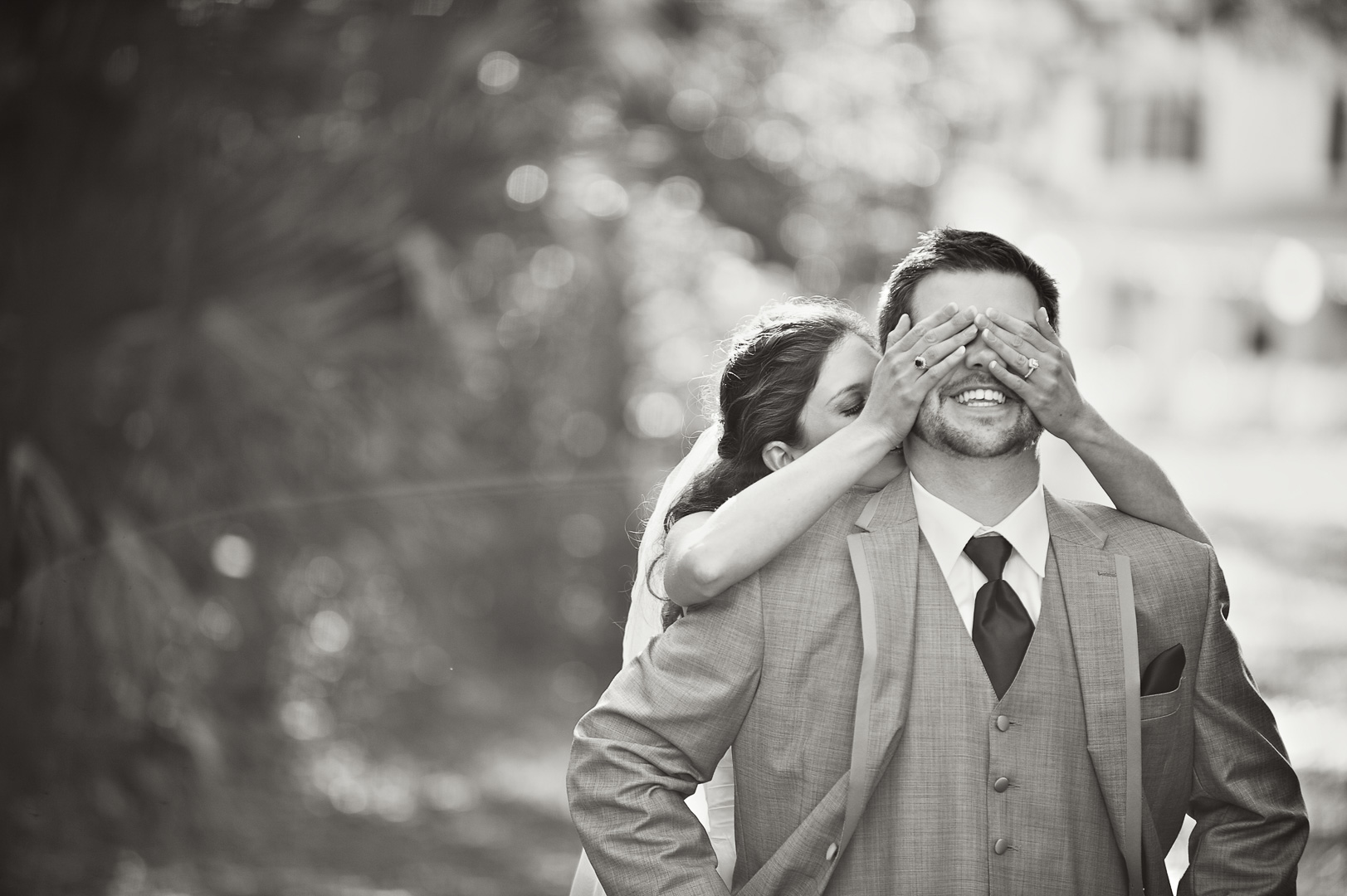 Person covering a man's eyes from behind, both smiling outdoors, blurred background, black and white photo.