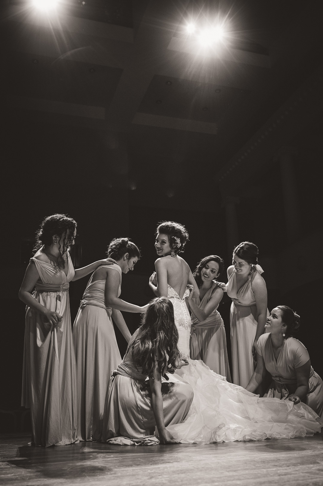 Seven women in elegant dresses gather around a woman in a wedding gown on stage, with bright lights overhead.