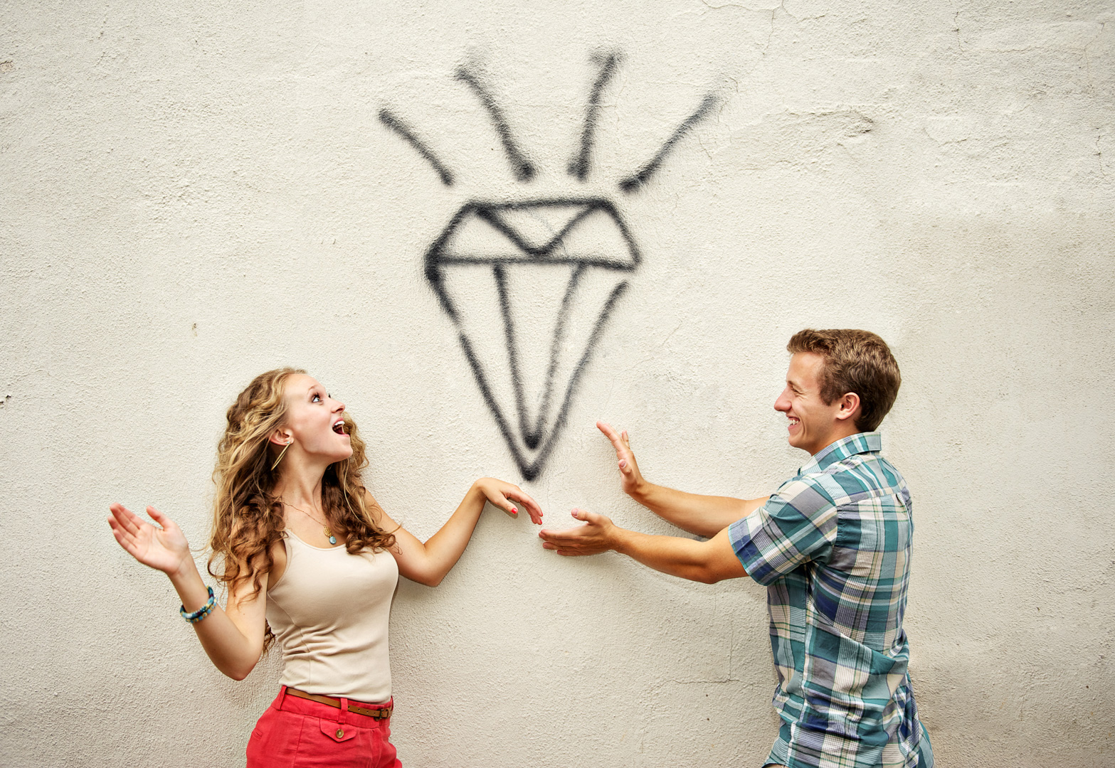 A woman and man smiling and gesturing towards a diamond drawing on a wall, with a plain background.
