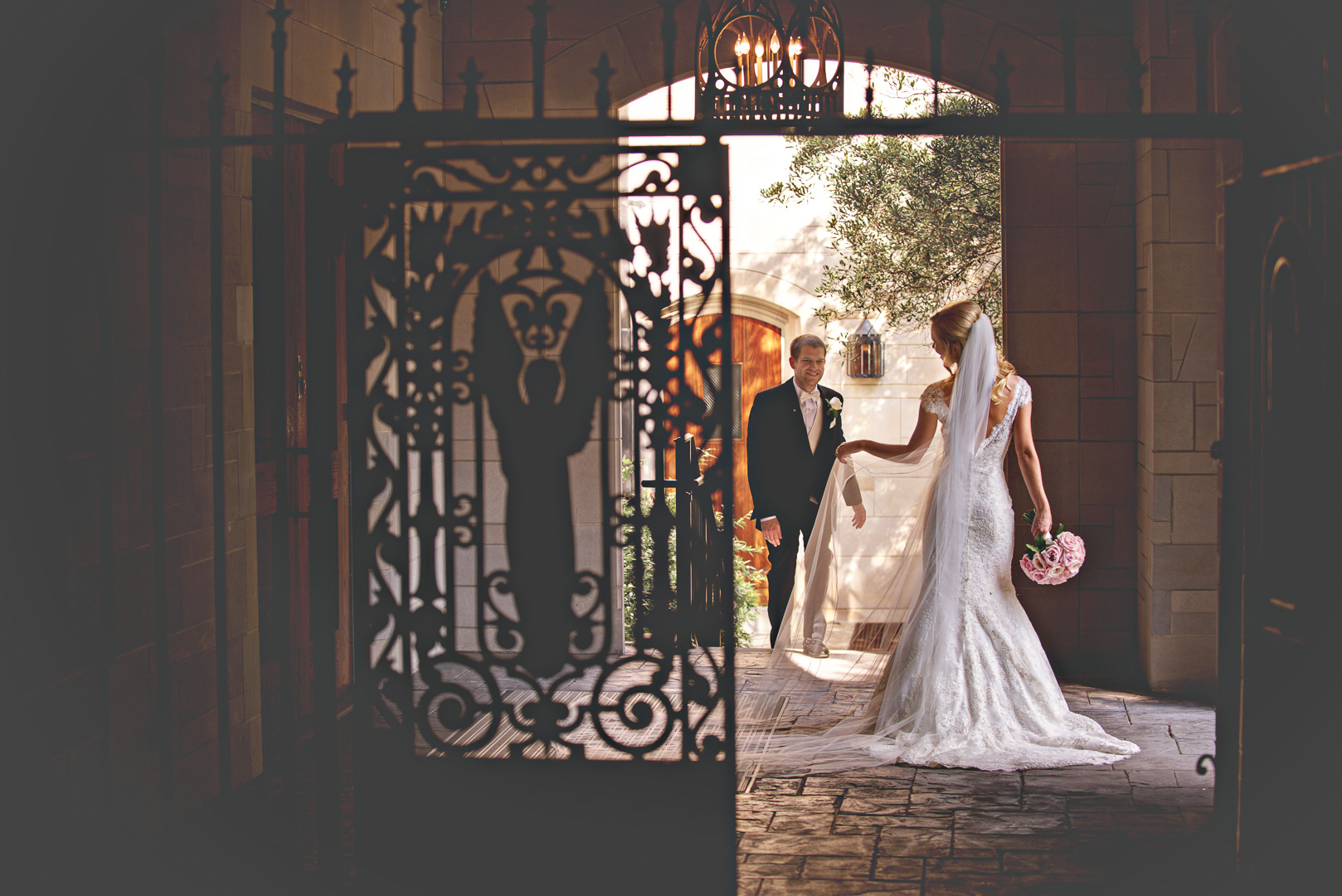 Bride and groom holding hands outside a building, with a decorative gate in the foreground, sunlight and trees in the background.