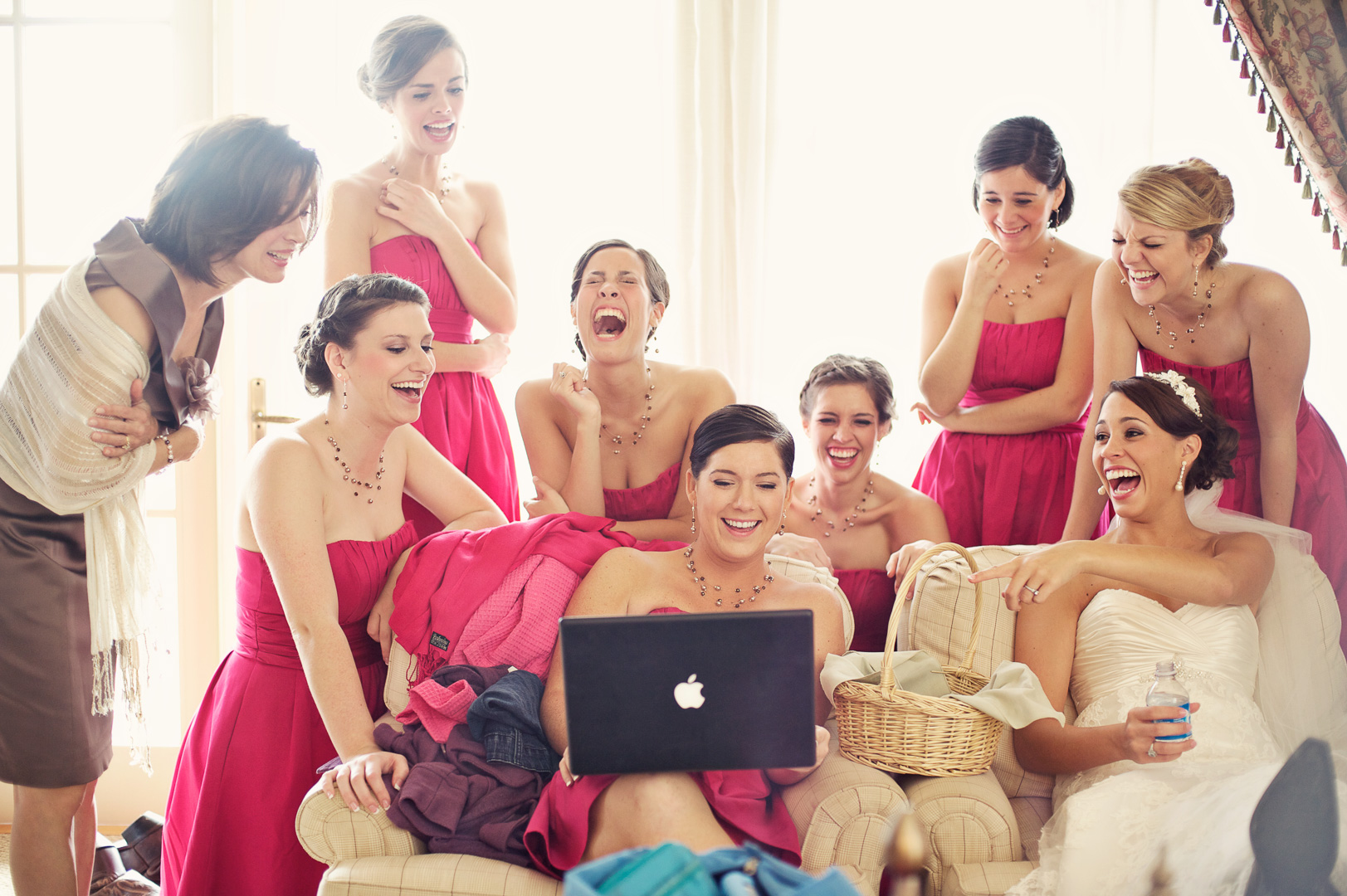 Group of women in pink dresses gathered around a woman with a laptop, laughing and smiling in a bright room.