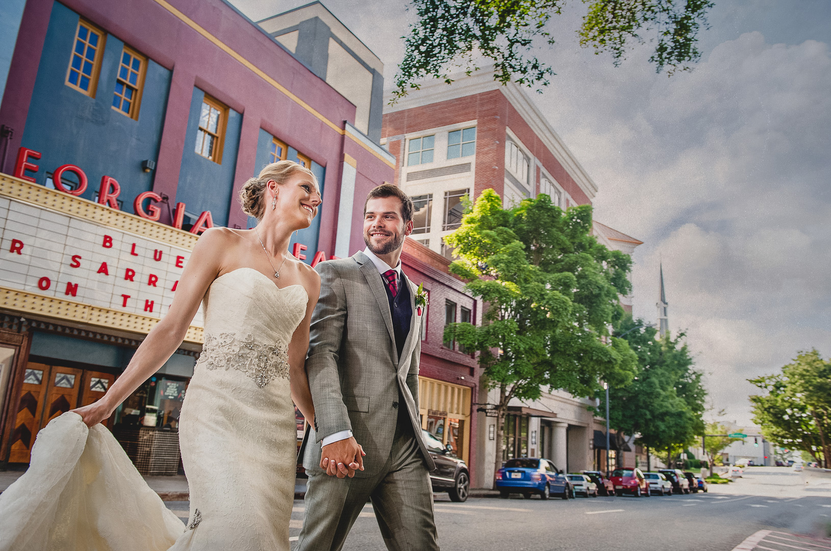 A bride and groom holding hands walking on a city street with colorful buildings and trees.
