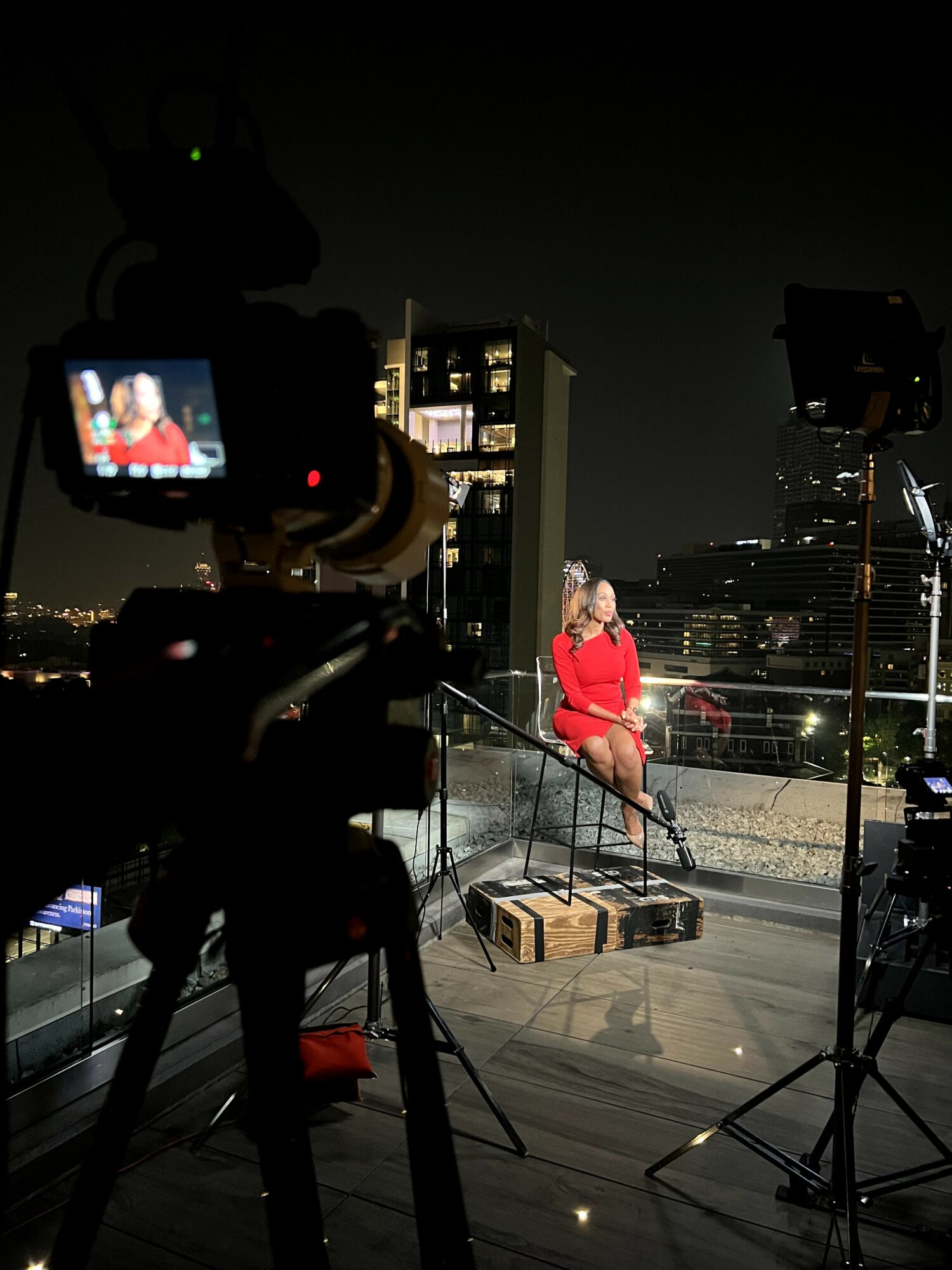 Person in red coat sitting on a box on a rooftop at night with city lights in background, surrounded by camera equipment.