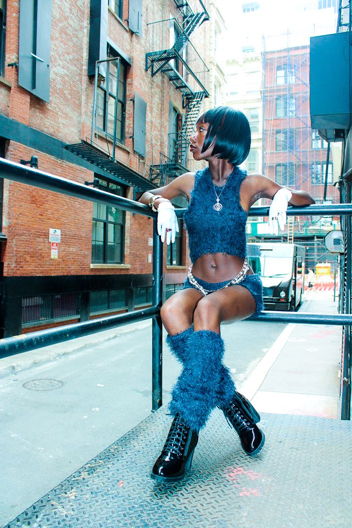 Person with short hair in a blue outfit and boots sitting on a railing in an urban alleyway.