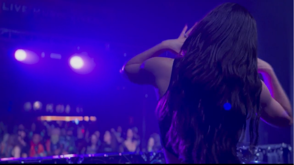 A woman with long dark hair is DJing at a concert with purple and blue stage lights, and a crowd in the background.