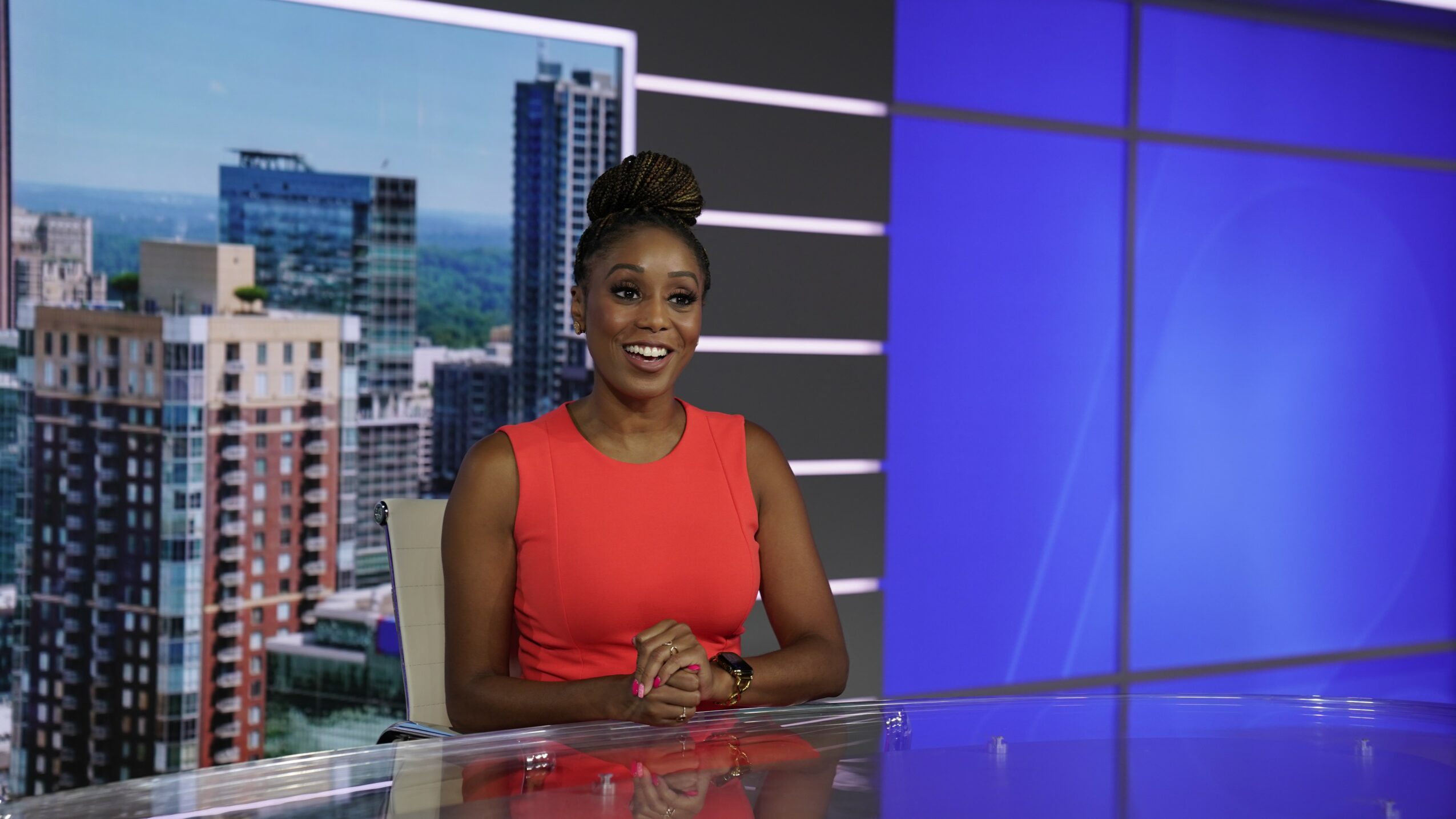 Woman in red sleeveless top sitting at a news desk with cityscape background, smiling, hands clasped, studio setting.