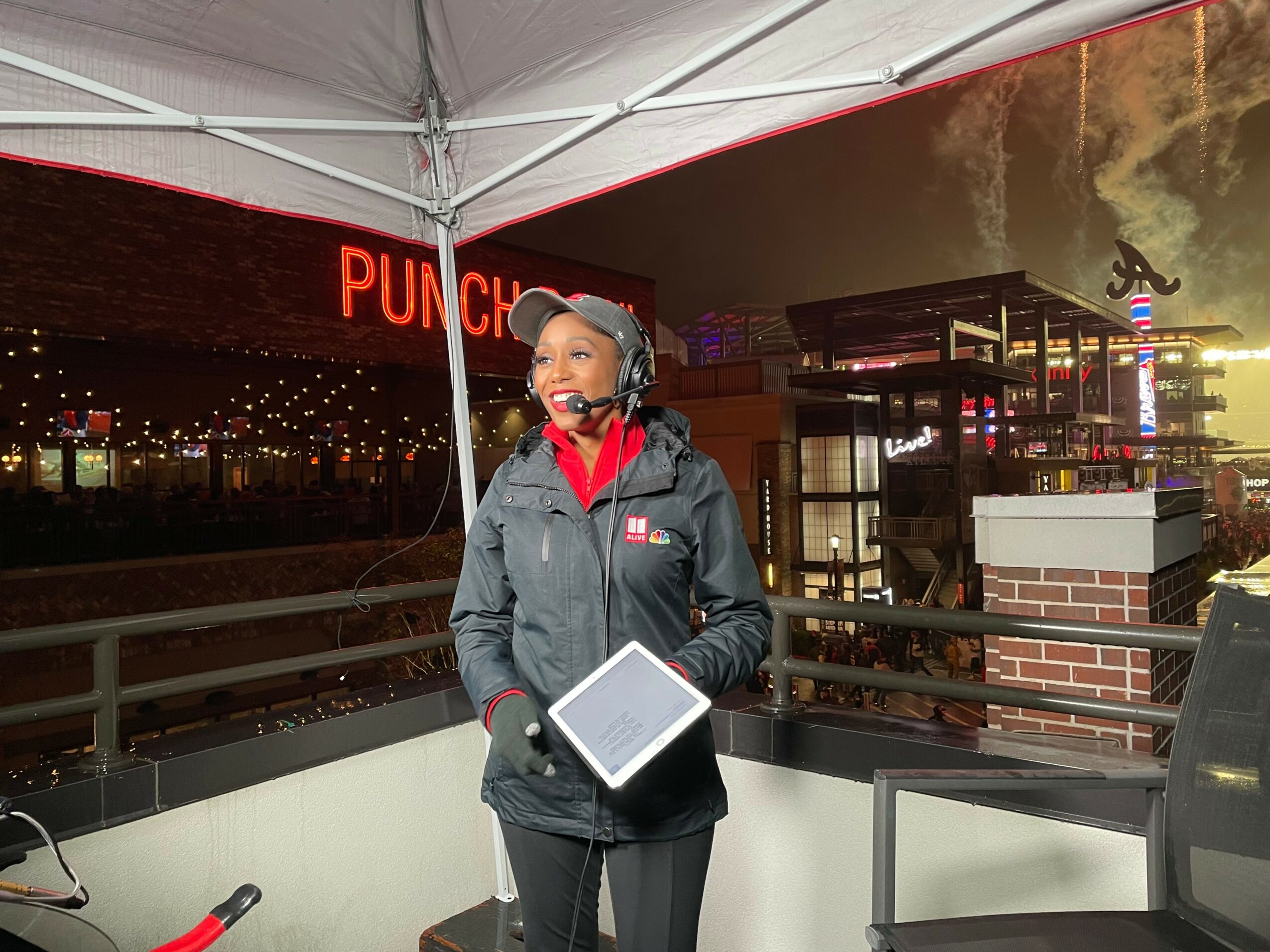 Woman wearing a headset and jacket holding a tablet, standing outdoors at night with city lights in background.