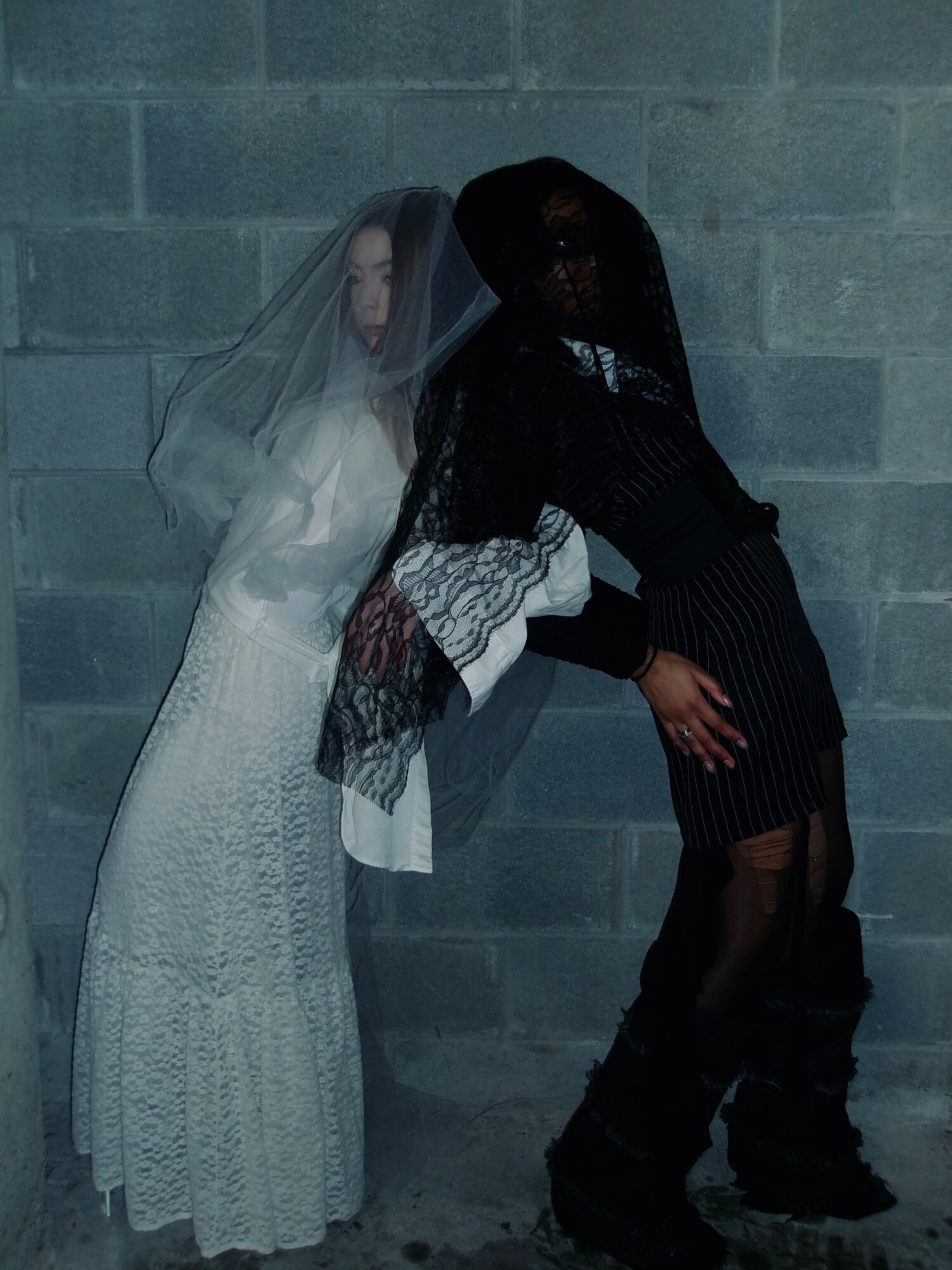 Two women in dark and light clothing with veils, standing against a gray brick wall, leaning towards each other.