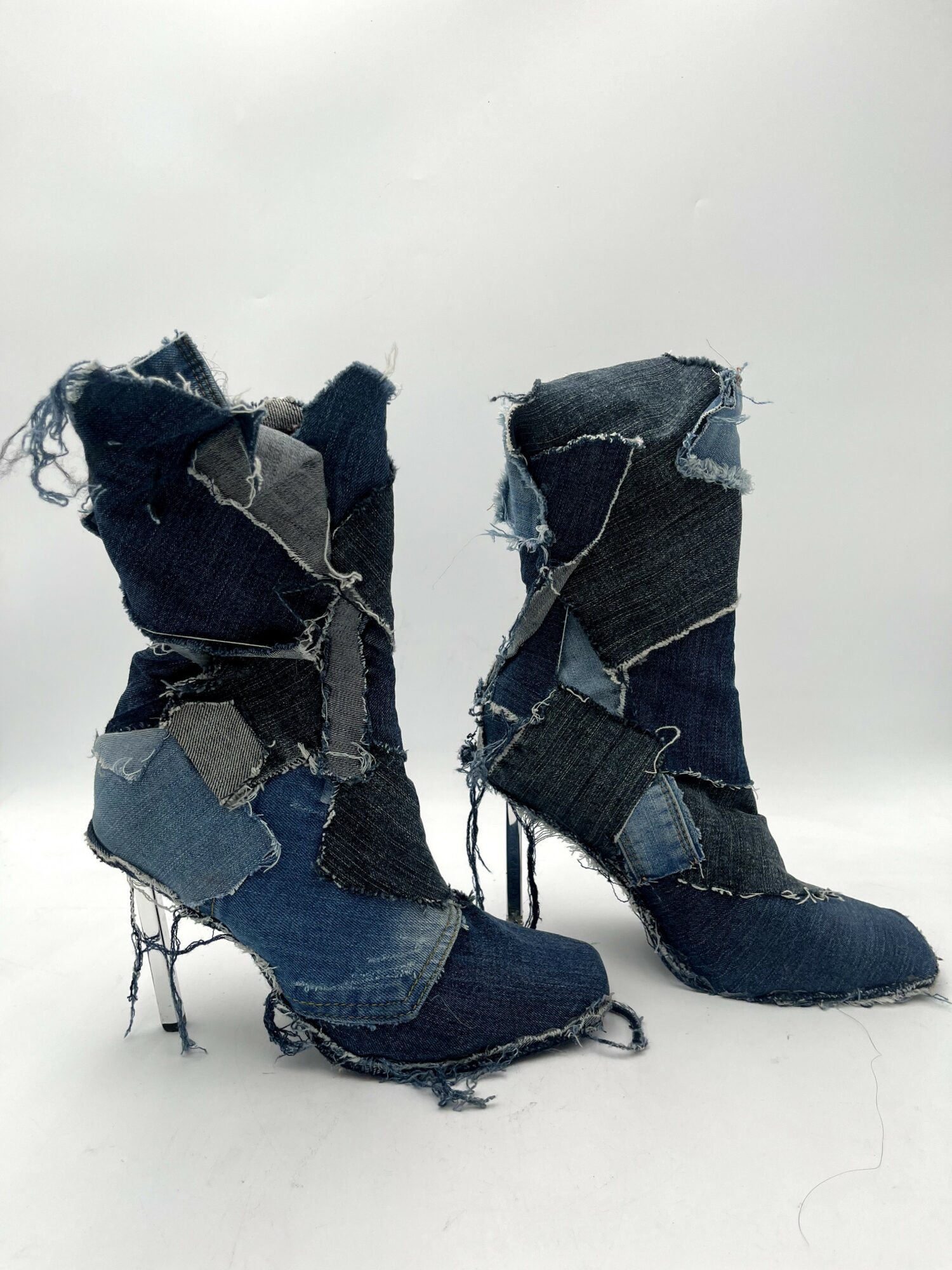 Pair of high-heeled boots made from torn denim fabric, with frayed edges and uneven patches, on a white background.