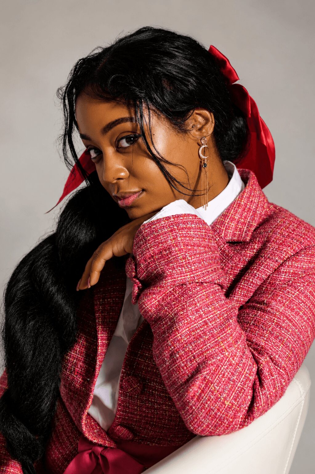 Young woman with long black hair, red bow, wearing a pink blazer and white shirt, sitting and looking at the camera.