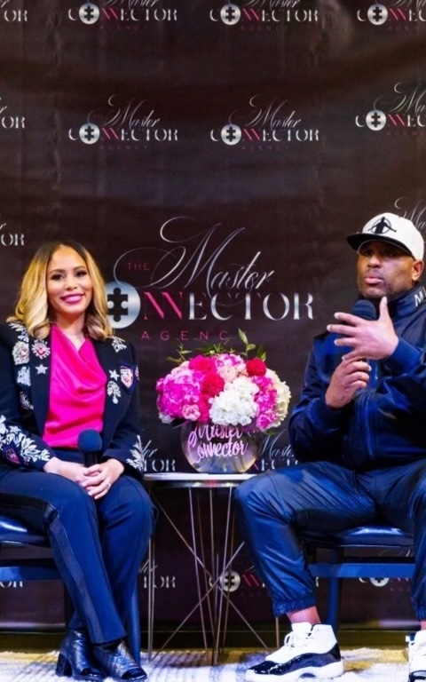 Two people sit on chairs with a table and pink flower arrangement between them, in front of a backdrop with text.