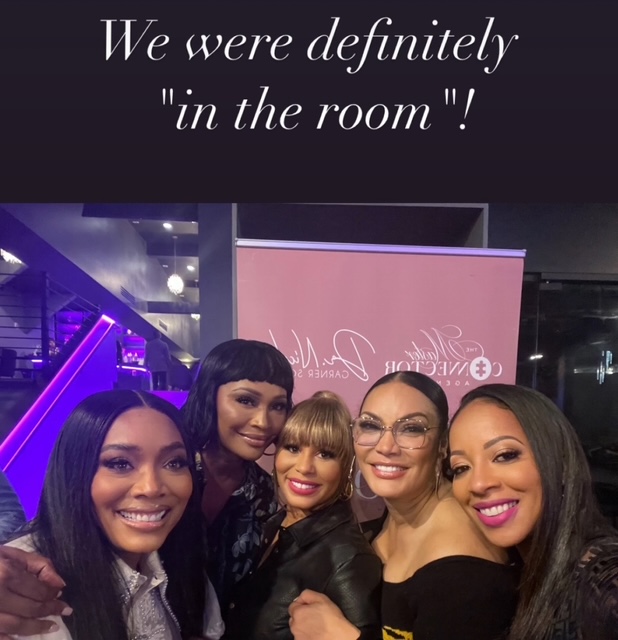 Four women smiling together indoors, with a pink banner and purple lighting in the background.
