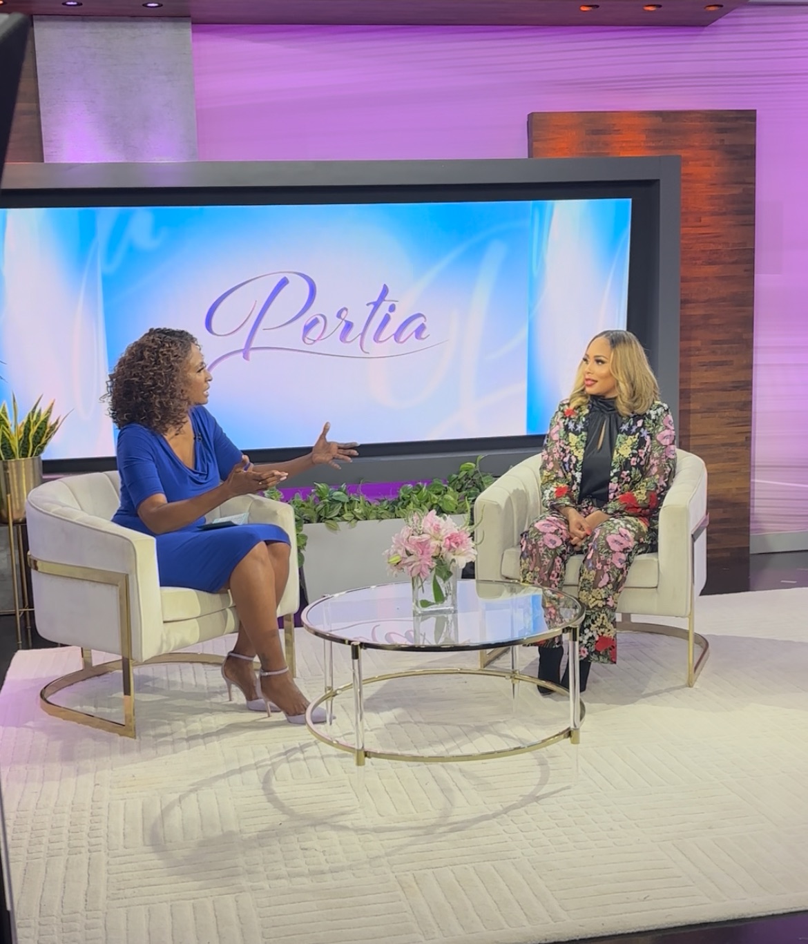 Two women sit on white armchairs in a TV studio, talking with a large screen behind them displaying the word 'Portia'.