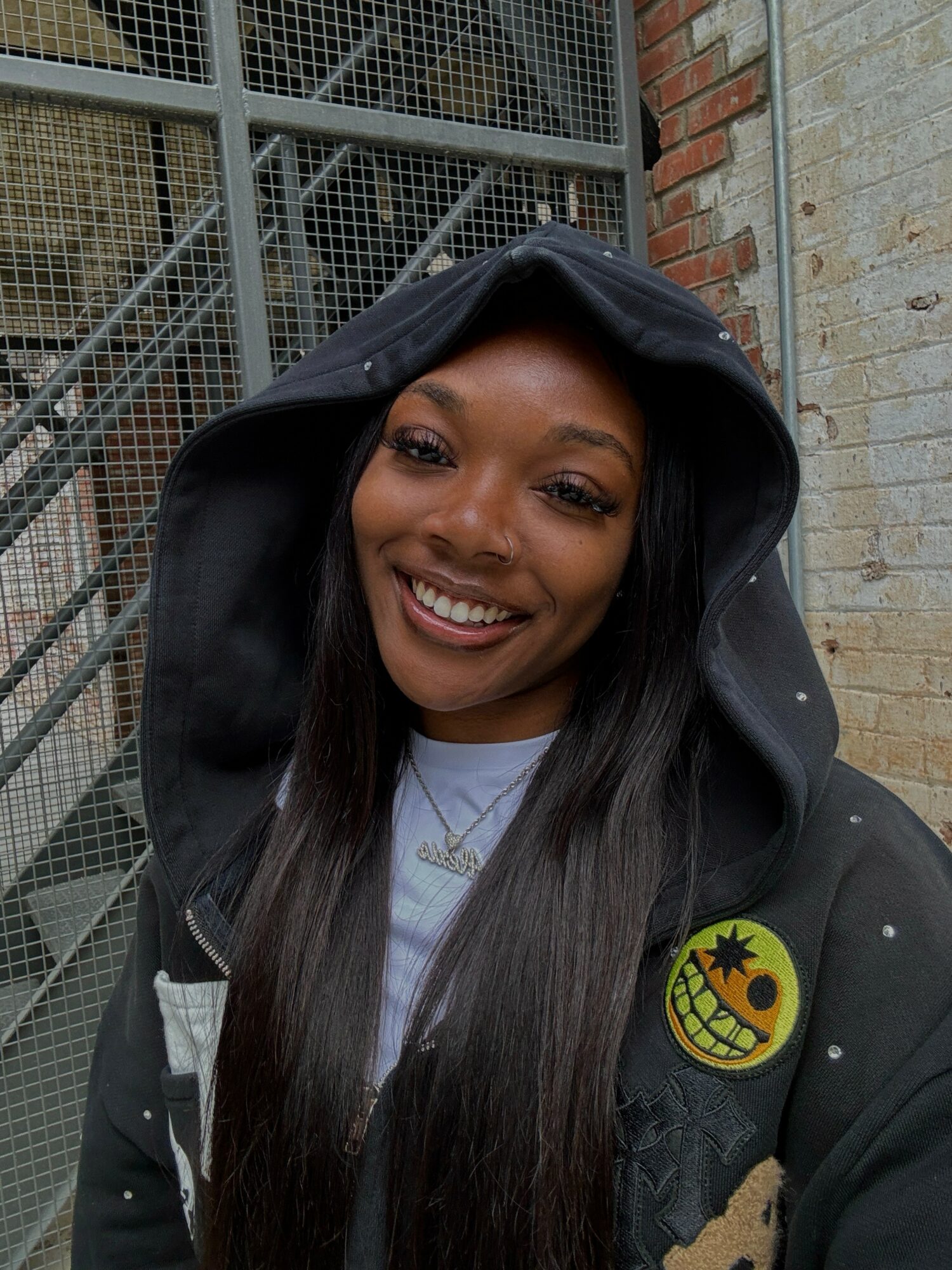 Smiling woman with long dark hair wearing a black hoodie outdoors, brick and metal fence background.