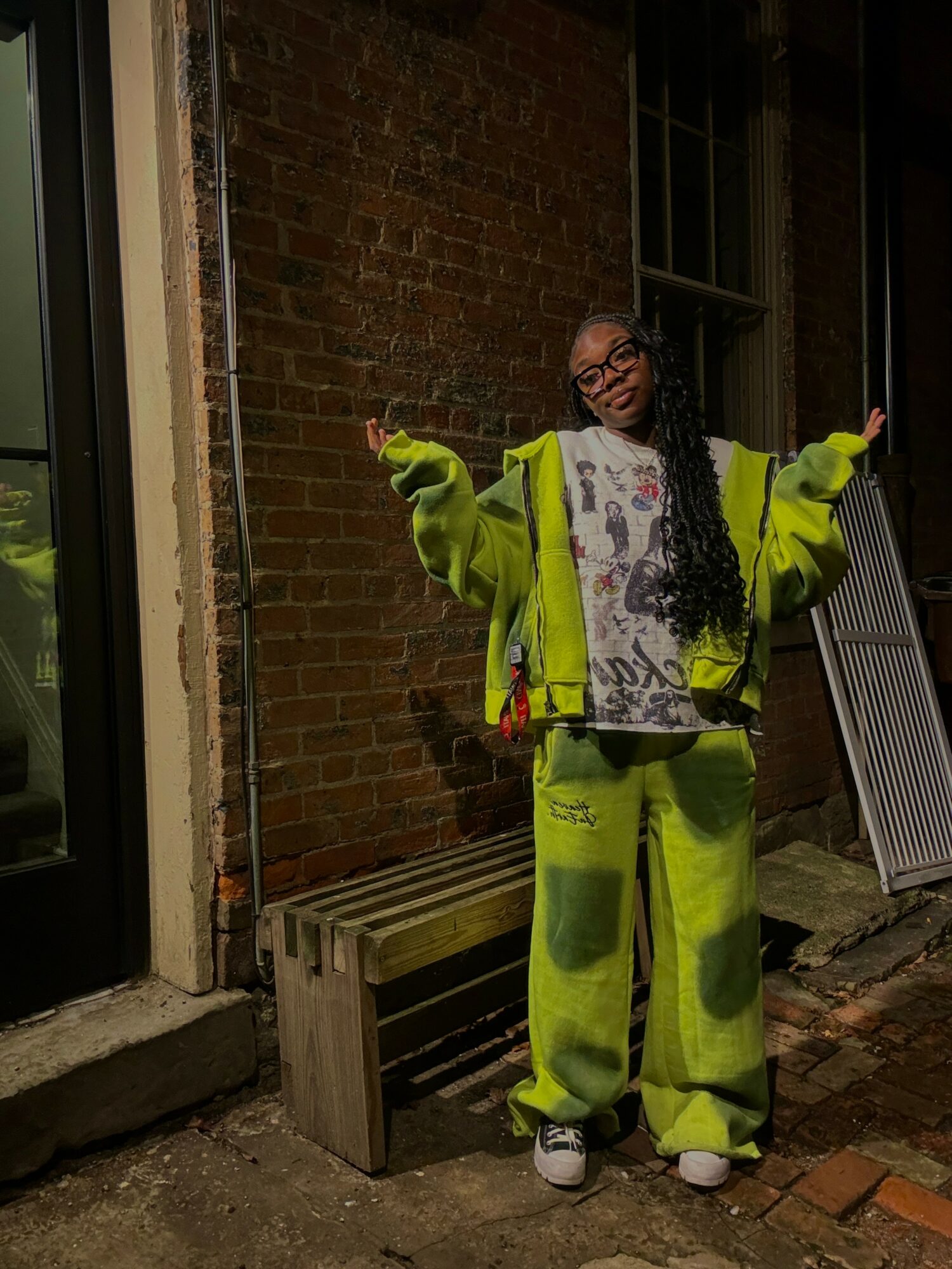 Person standing outdoors near brick wall, smiling, wearing bright yellow-green oversized jacket and pants, with arms outstretched.
