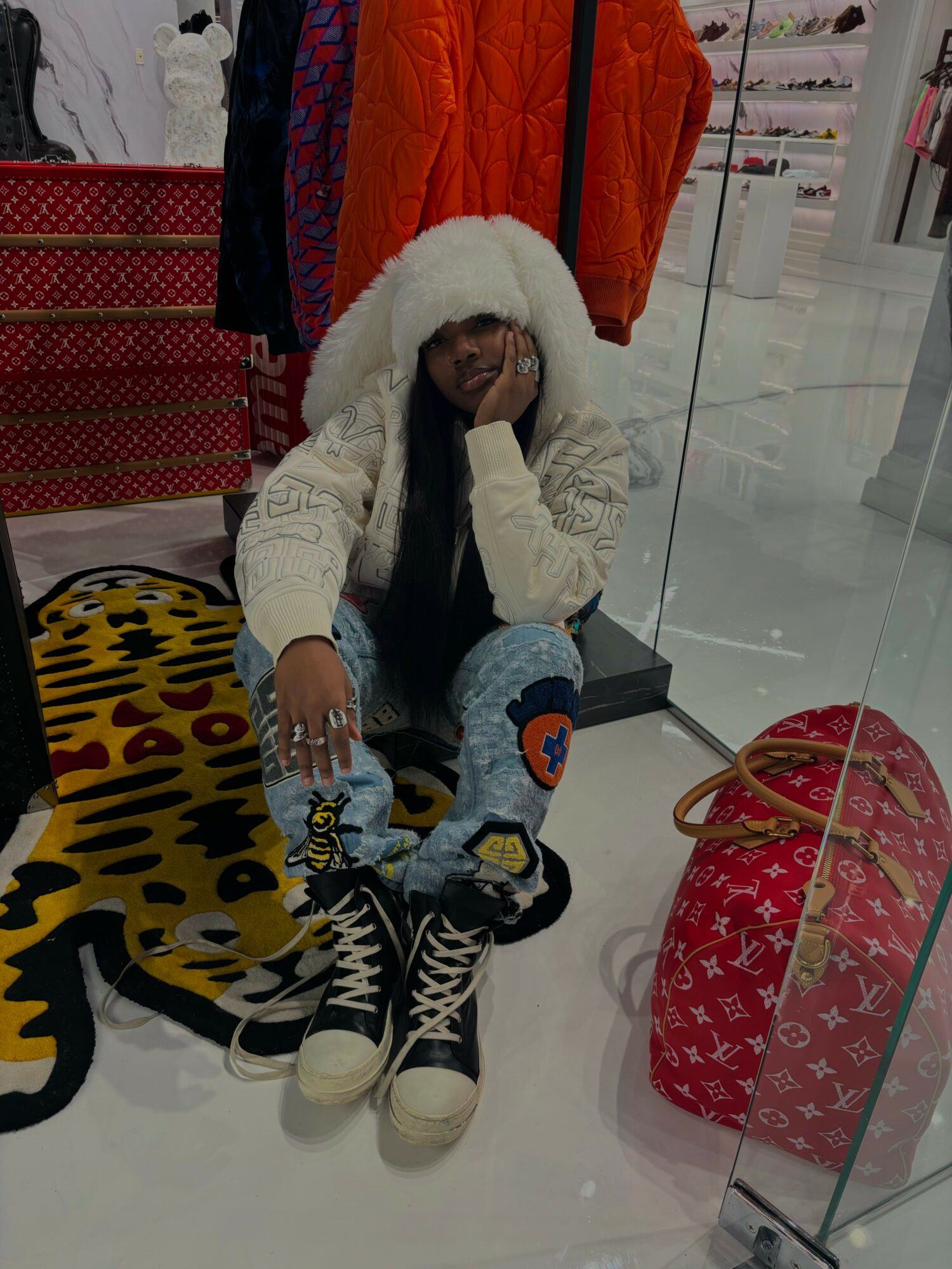 Child sitting on the floor with a white furry hood, surrounded by shopping bags, in a store with glass walls.