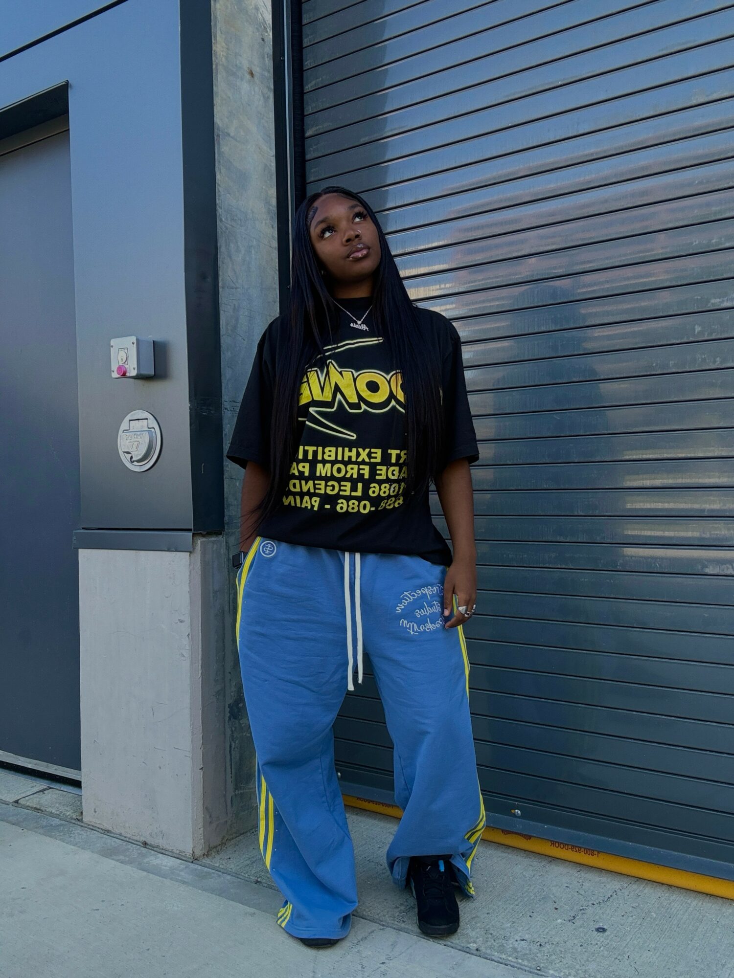 Young woman with long braids standing against a gray wall and metal shutter, wearing a black graphic T-shirt and blue pants.