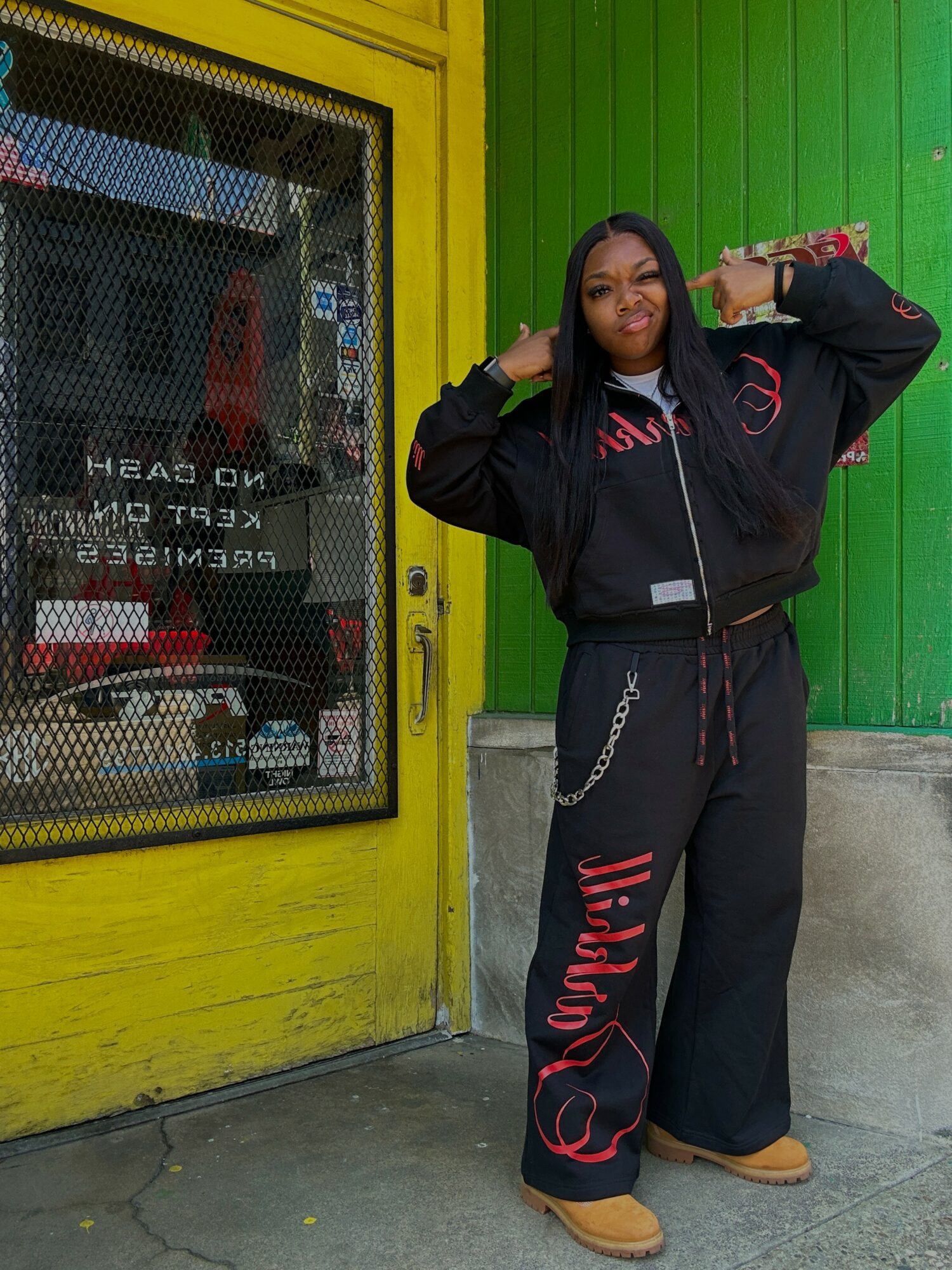 Woman standing outside in front of a yellow and green wall, wearing a black hoodie and pants, with one hand on her head.