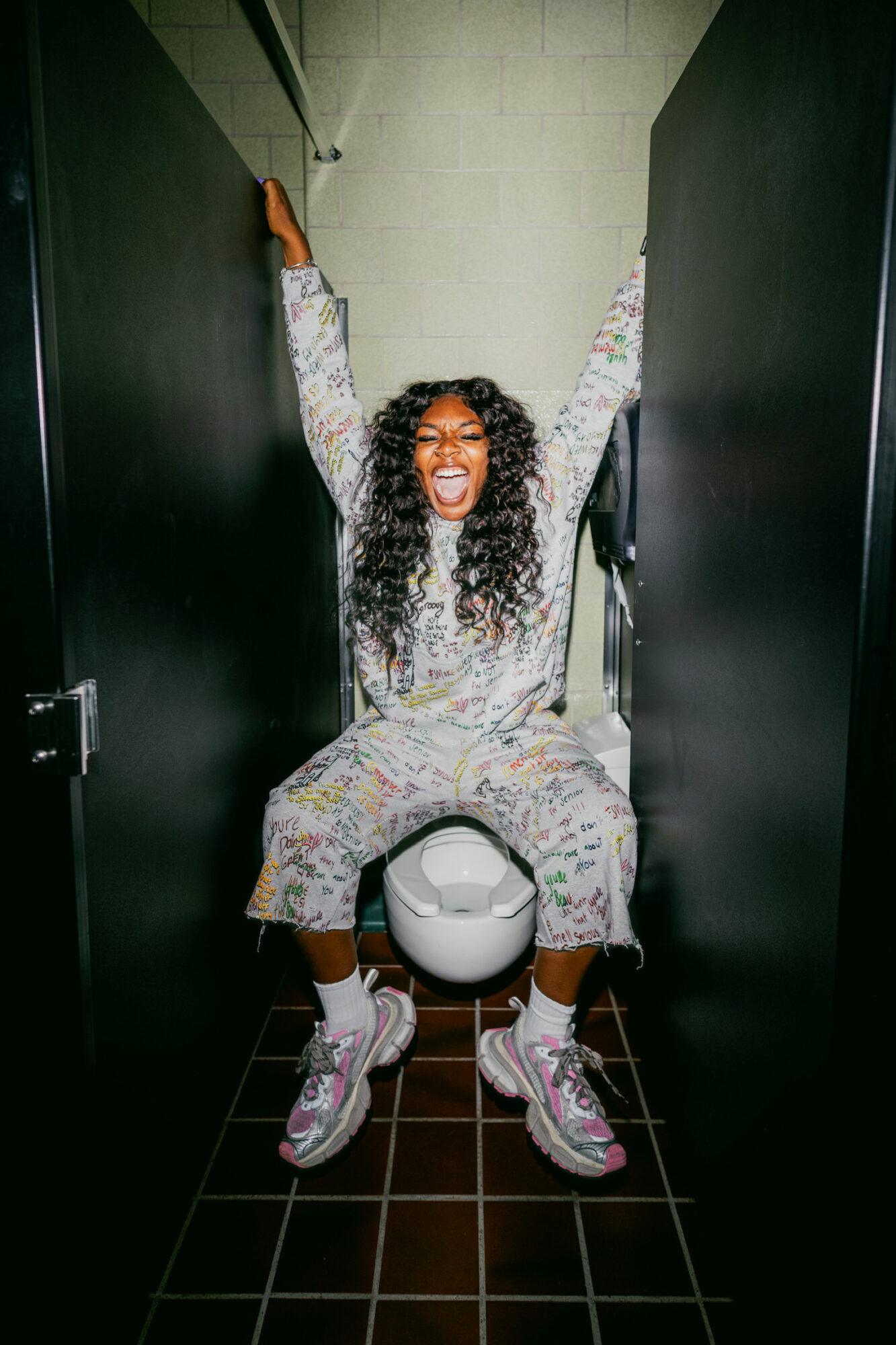 Person with curly hair smiling, sitting on toilet in a narrow bathroom, arms raised, wearing colorful pajamas and sneakers.