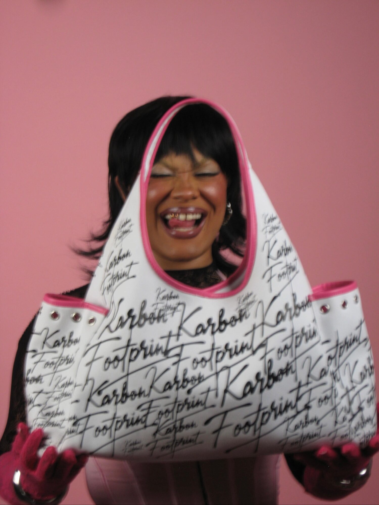 Person with black hair and pink gloves holding a large white bag with black handwritten text, smiling against a pink background.