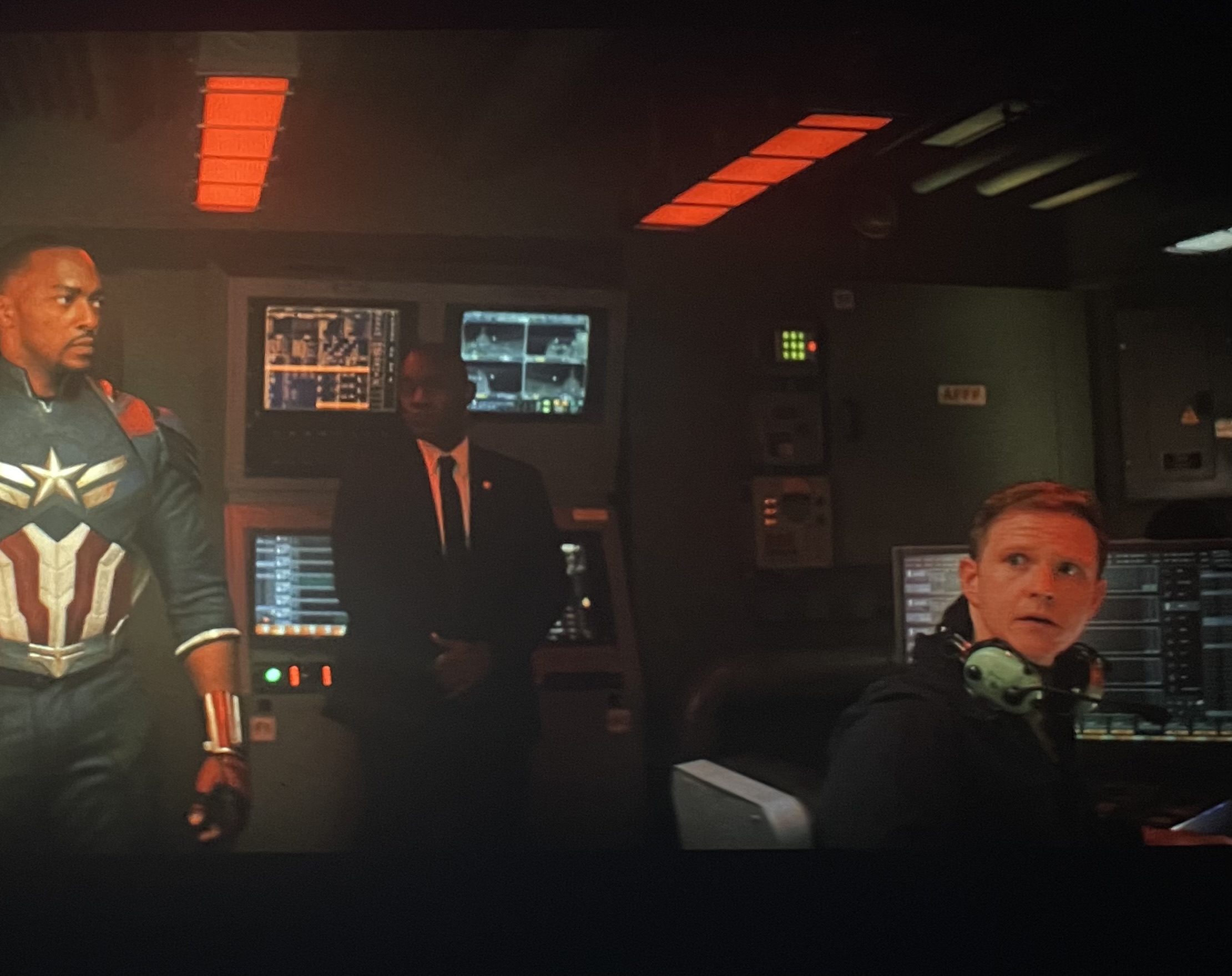 Three people in a control room with screens and equipment, one in a superhero costume, one in a suit, and one at a computer.
