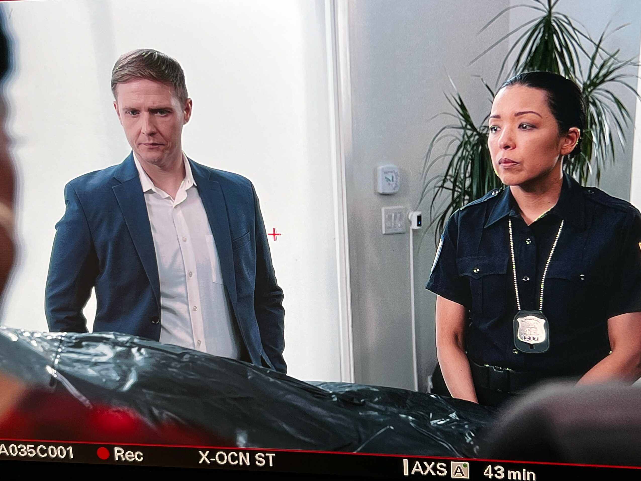 Two people, a man in a suit and a woman in a police uniform, stand indoors near a car, with plants in the background.