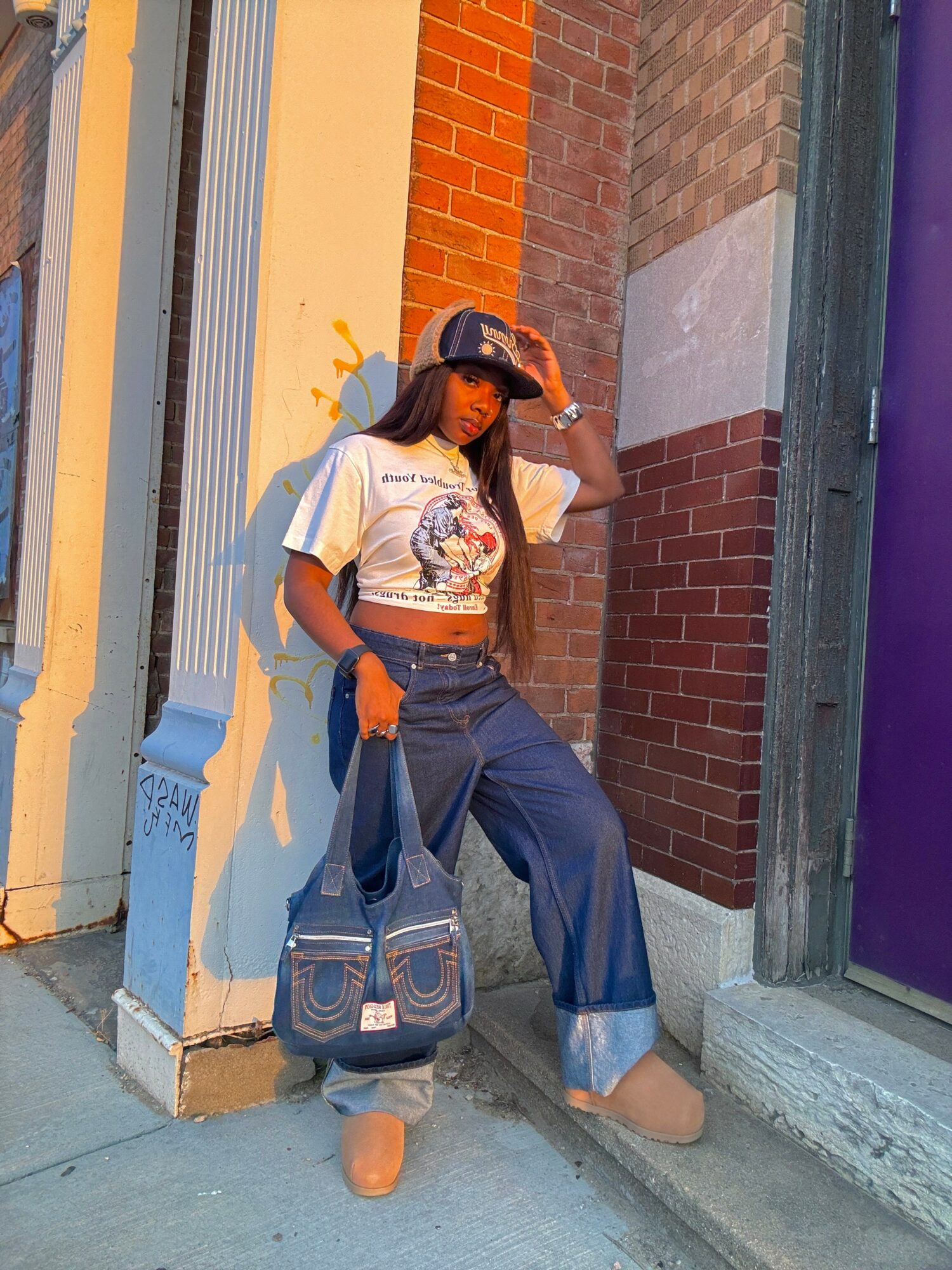 Young woman with long hair wearing a white graphic t-shirt, wide-leg jeans, and tan boots, standing against a colorful urban wall, holding a bag, and shielding her eyes from the sun.