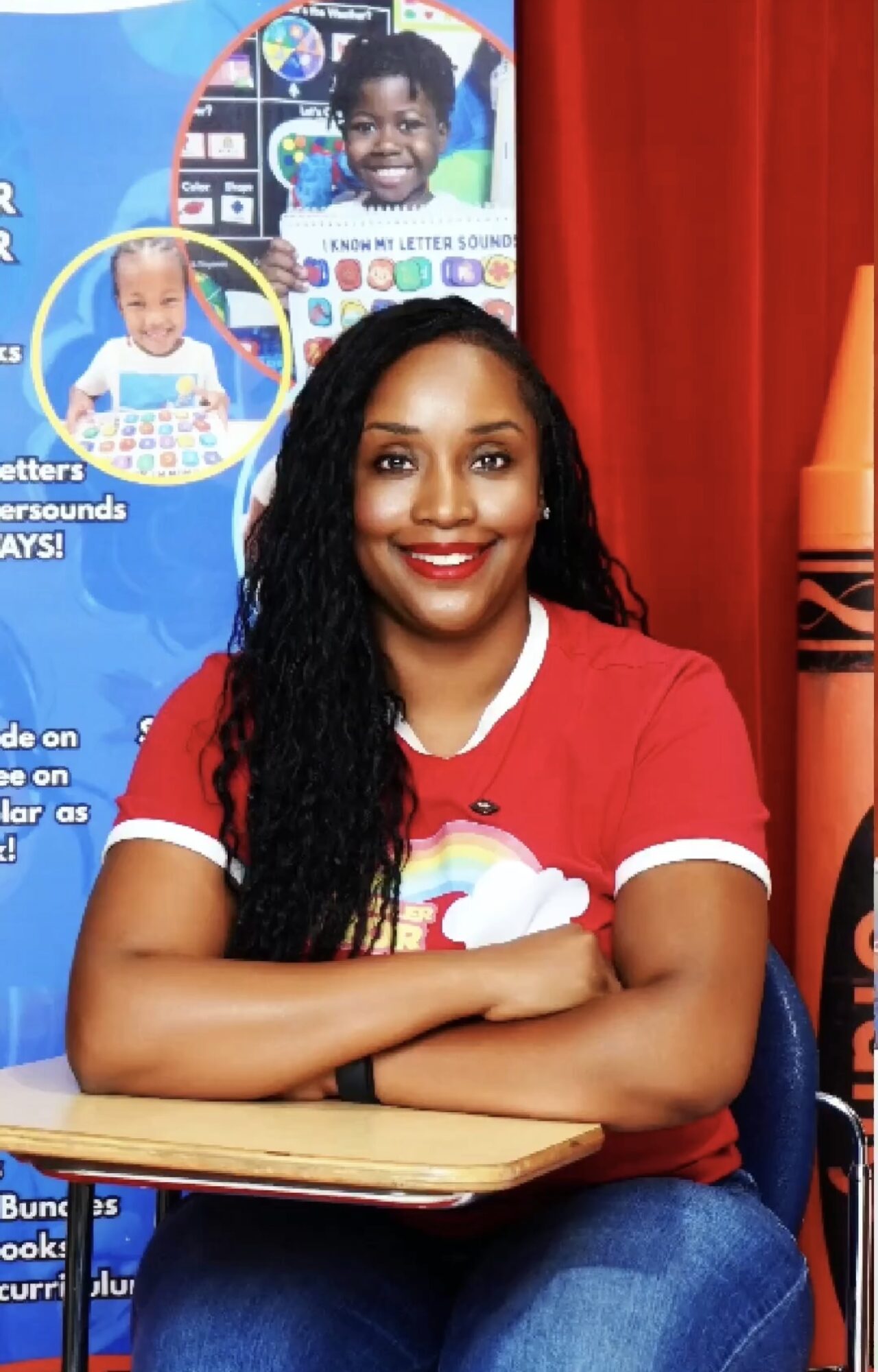 Smiling woman with long curly hair sitting at a table in front of educational posters, wearing a red shirt with a rainbow design.