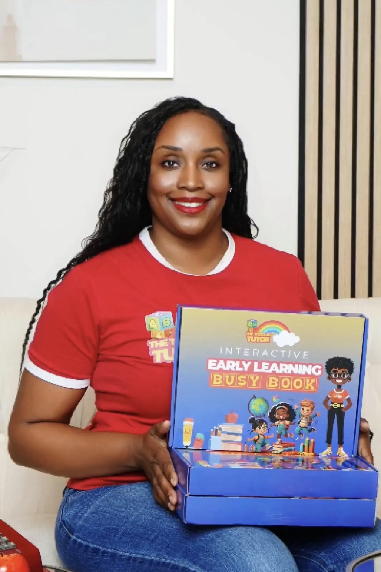 Woman with long curly hair wearing a red shirt holding a blue box with children and educational graphics inside.