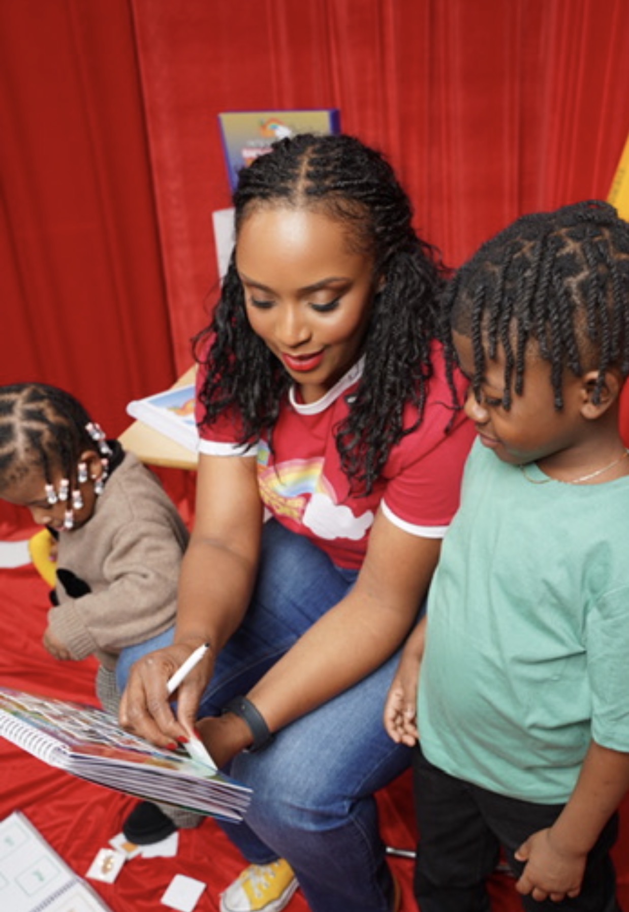 Woman with curly hair reading a book to two children, one with braided hair and the other with dreadlocks, in front of red curtains.