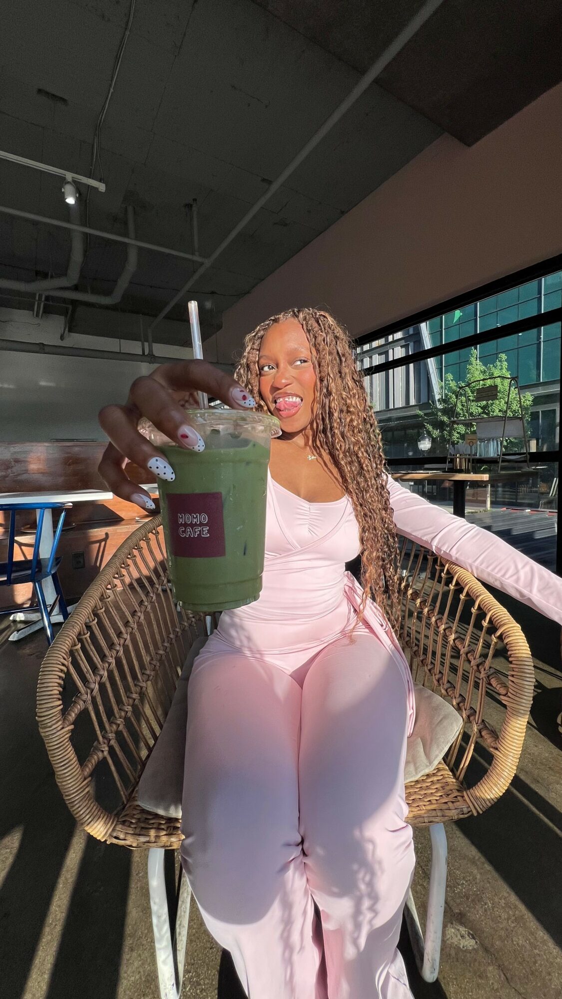 Woman with long curly hair sitting in a wicker chair, holding a green drink, smiling, in a modern indoor setting with large windows.