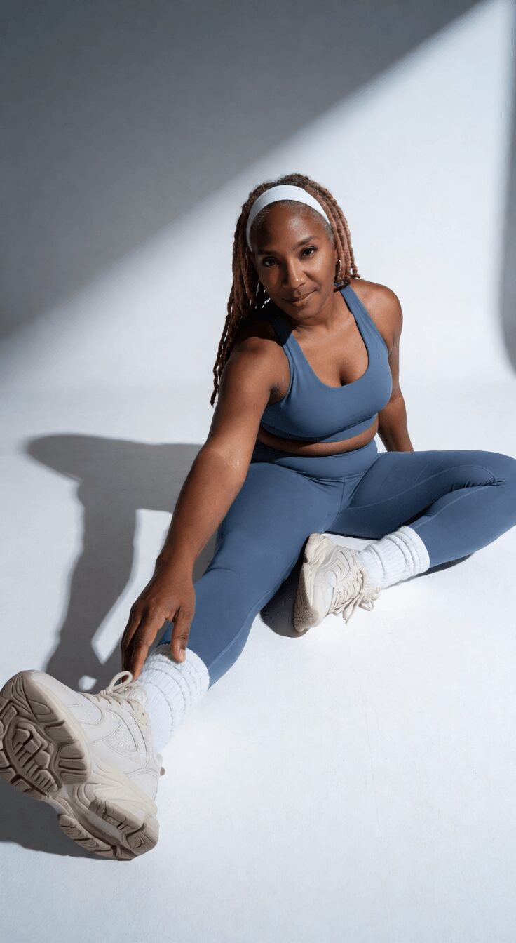 Woman in blue workout outfit sitting on the floor with legs extended, white sneakers, and headphones, against a plain background.