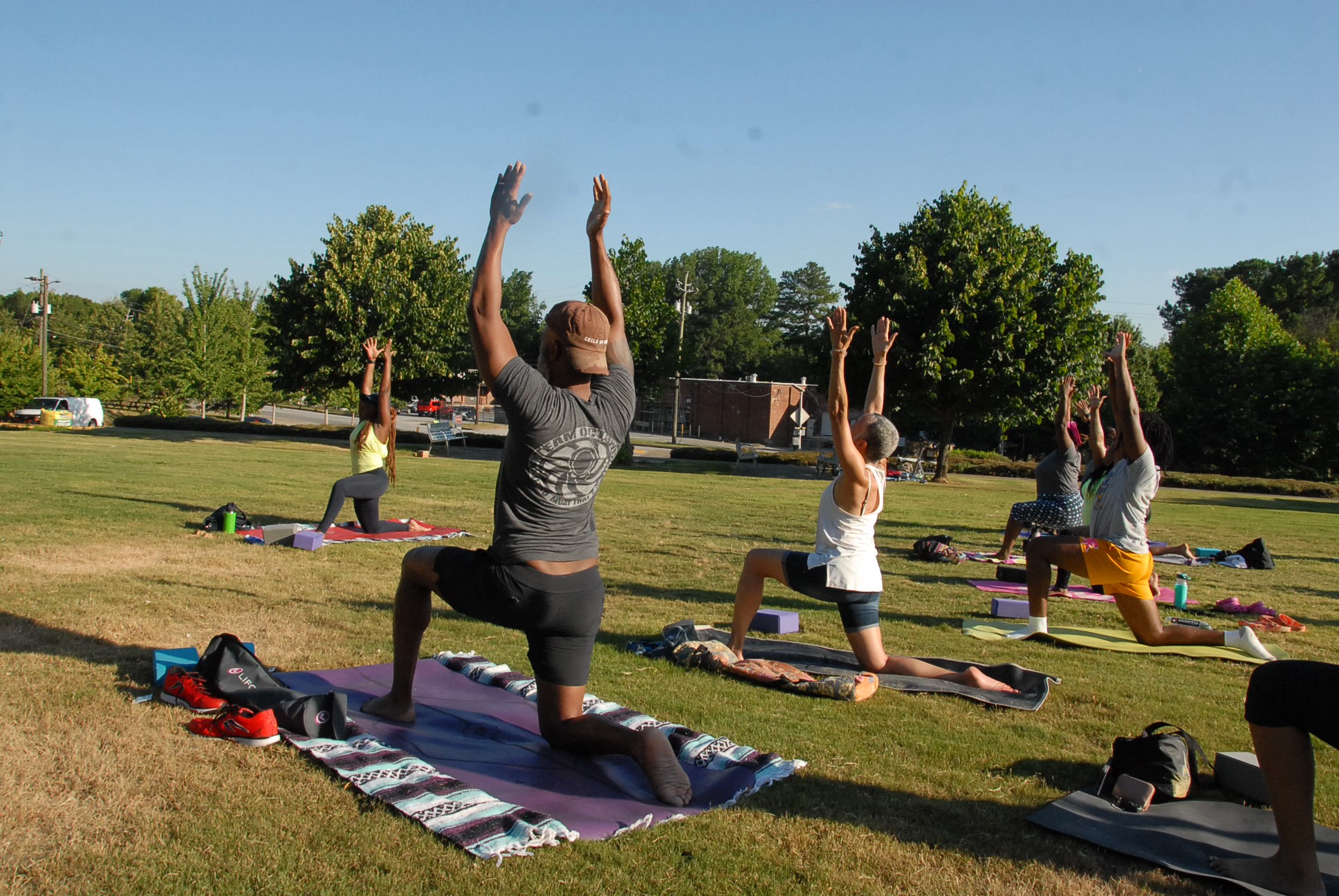 Group of people practicing yoga outdoors on mats in a park with trees and blue sky.