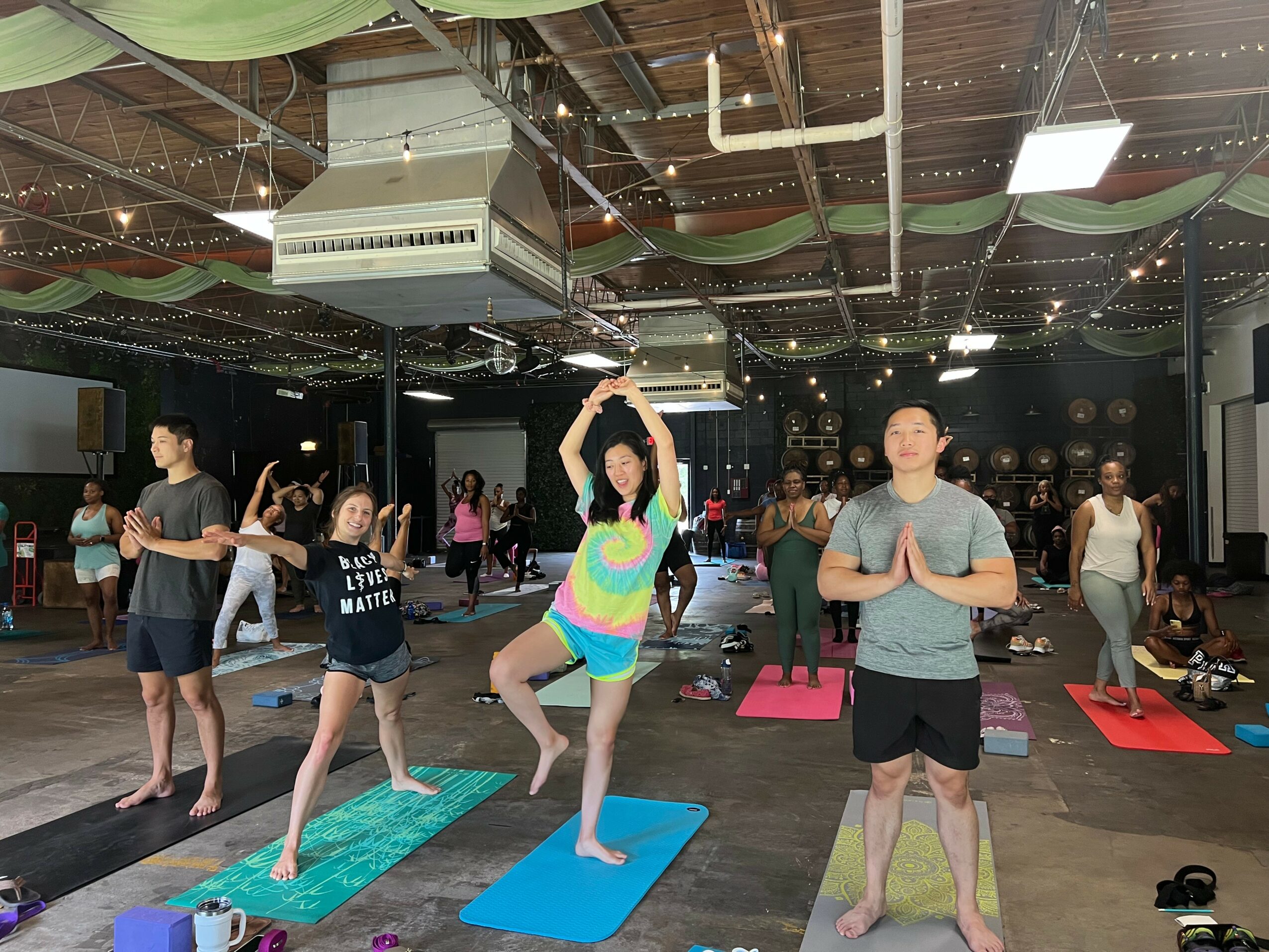 Group of people practicing yoga on mats in a spacious indoor studio with string lights and ceiling decorations.