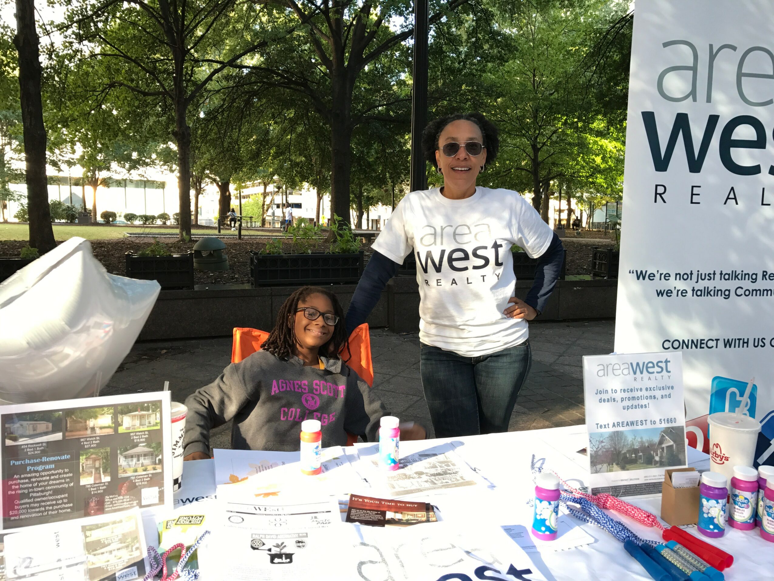 Two women at an outdoor booth with trees in the background, one sitting and one standing, smiling, with promotional materials on the table.