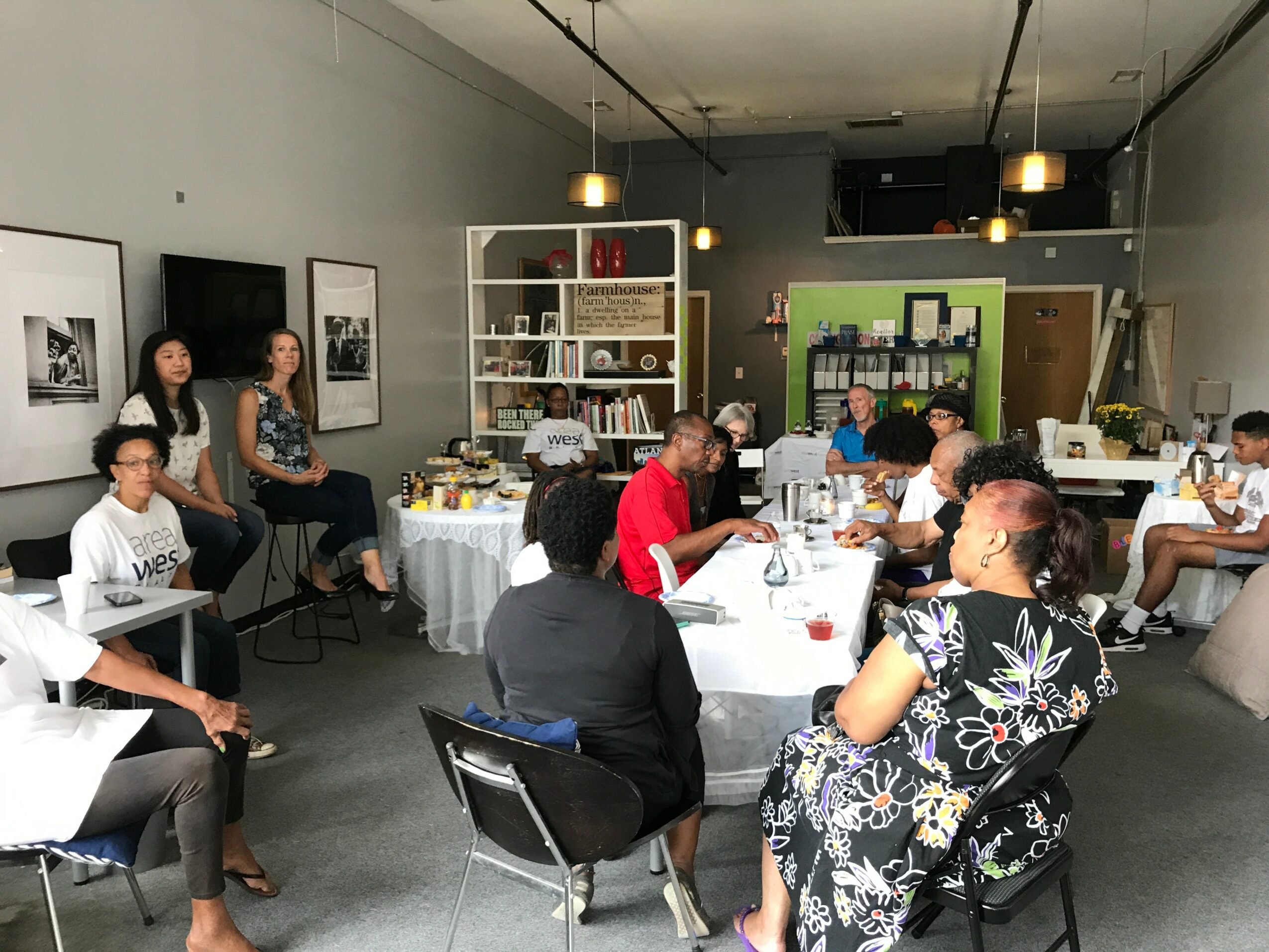 People seated around a table in a room with shelves, framed pictures, and a green wall, engaged in discussion.