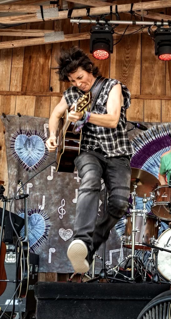 Person playing guitar on stage with colorful decorations and drums, wearing a checkered shirt and jeans.
