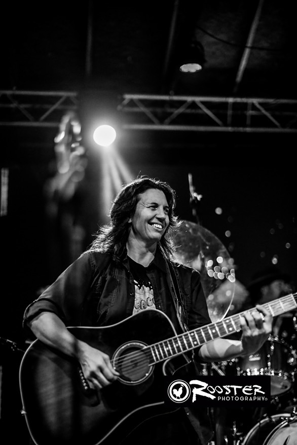 Woman playing guitar and smiling on stage with bright lights and equipment in background.