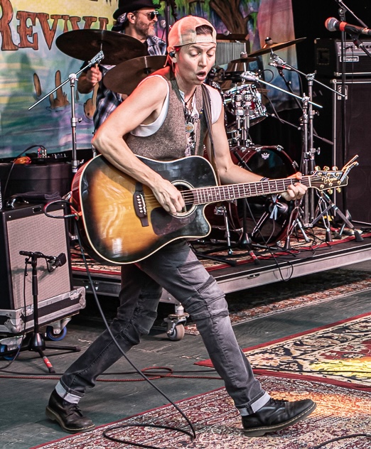 Musician playing an acoustic guitar on stage with drums and a colorful backdrop, wearing a cap and casual clothing.