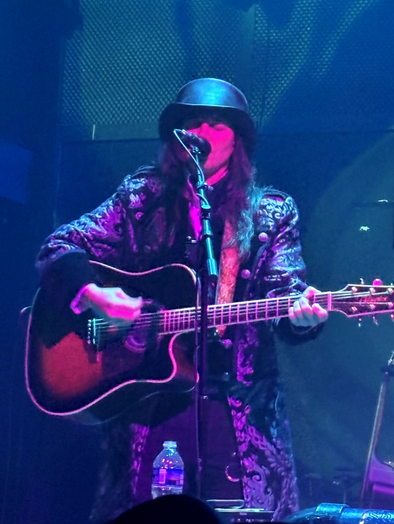 Person wearing a hat and patterned jacket playing an acoustic guitar on stage with a microphone, water bottle nearby.