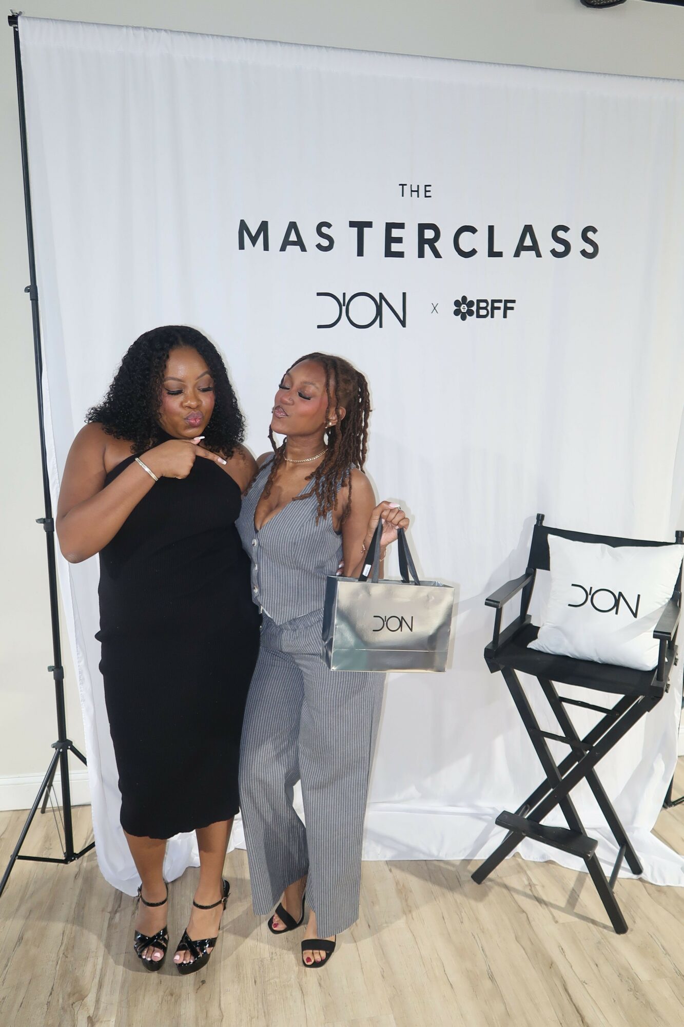 Two women standing in front of a white backdrop with black text, one holding a white bag, both smiling.
