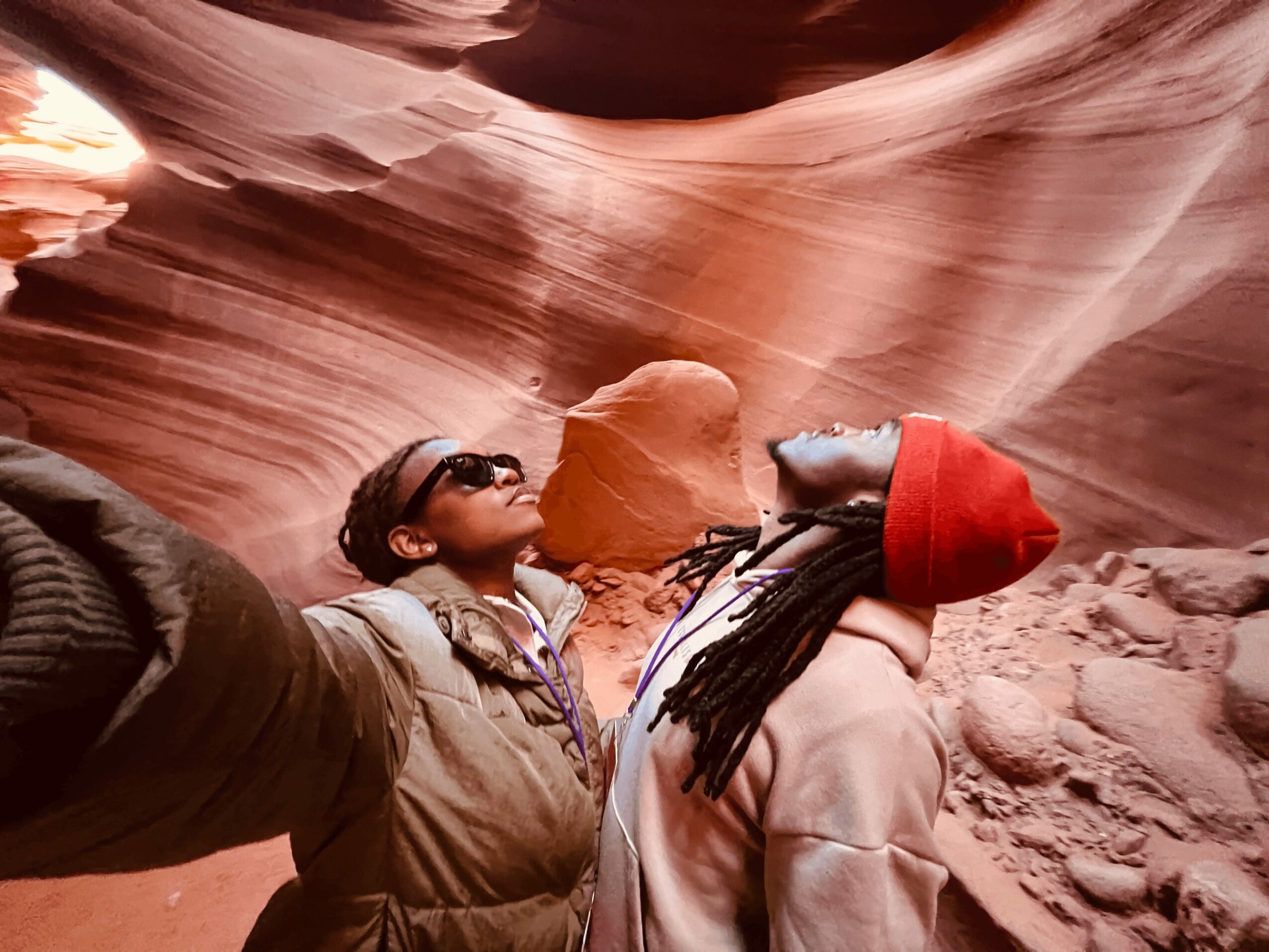 Two people in warm clothing and hats taking a selfie inside a narrow, colorful canyon with smooth, layered rock walls.